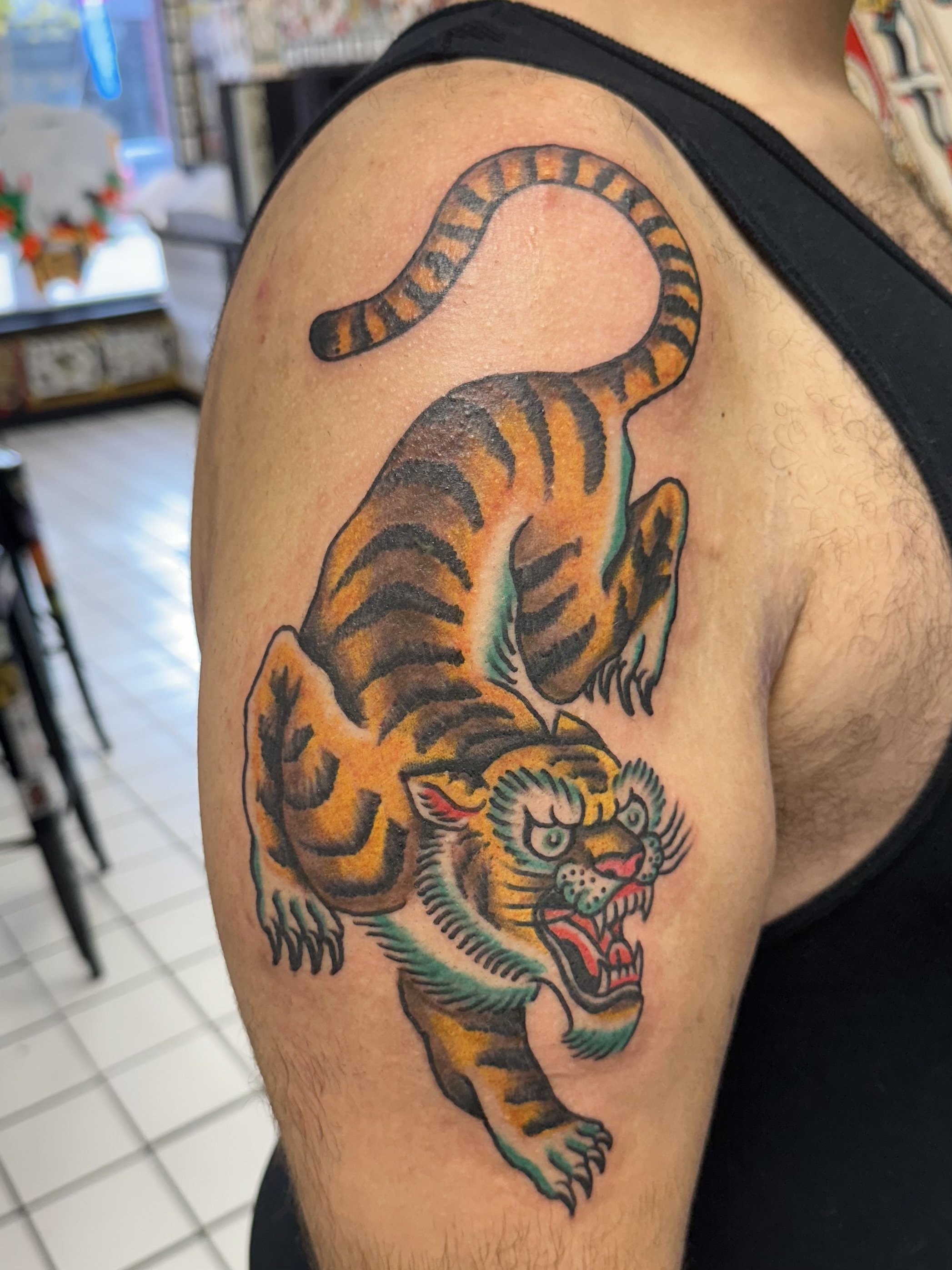 Colorful tattoo of a tiger on a person's upper arm, featuring bold black outlines and orange, green, and red hues.