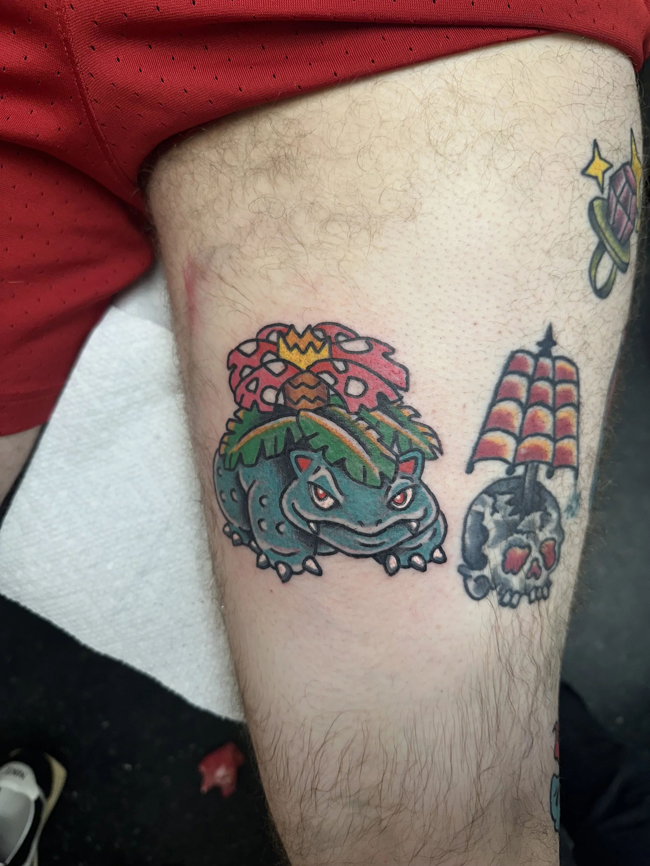 Tattooed tattoos on a person's thigh, including a Coffin with red, black, and purple colors, and a dragon with a blue face and green tongue, beneath the thigh of someone wearing red athletic clothing.