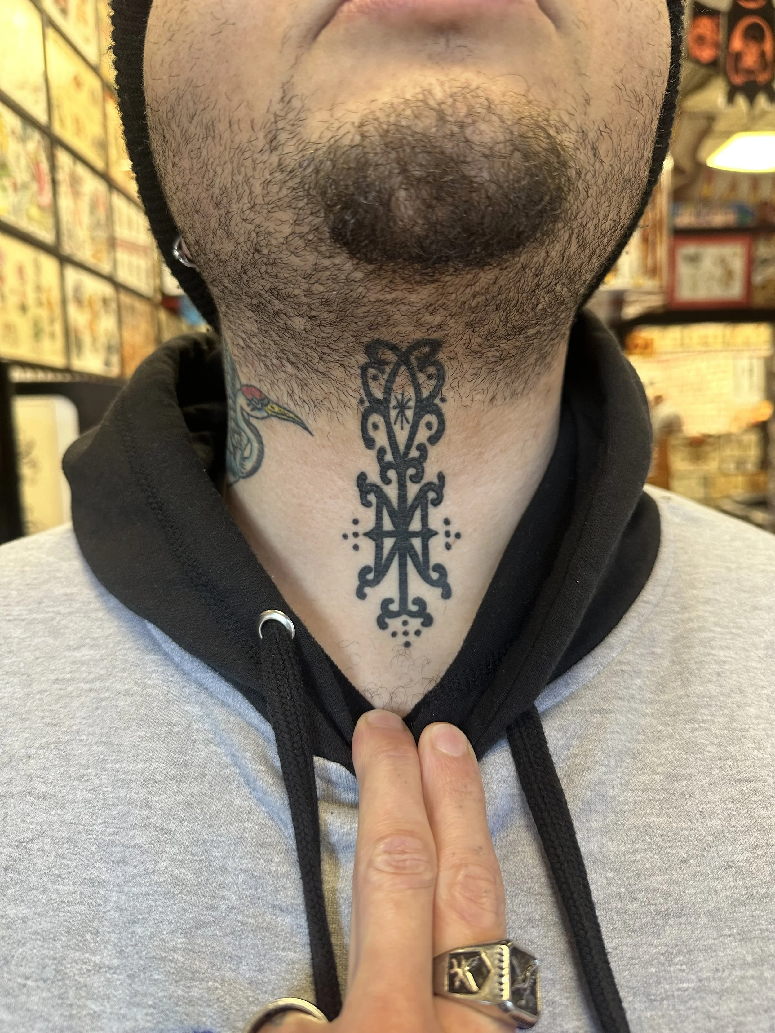 Close-up of a person's chin with facial hair, tattoos on the neck, and a hand with rings pointing toward the chin. The person is wearing a hoodie with a grey body and black hood.