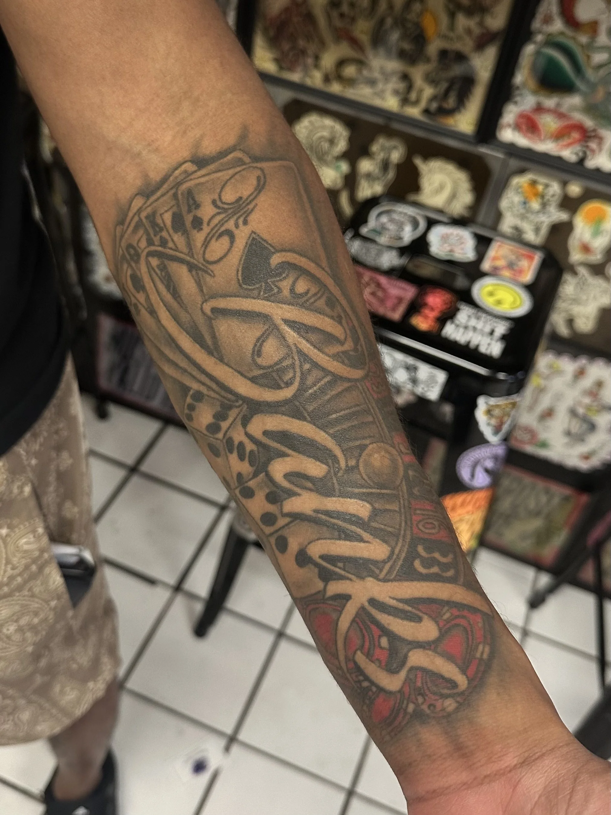 A person's arm with a large tattoo of a snake and playing cards, in an indoor setting with a display of various stickers or patches in the background.