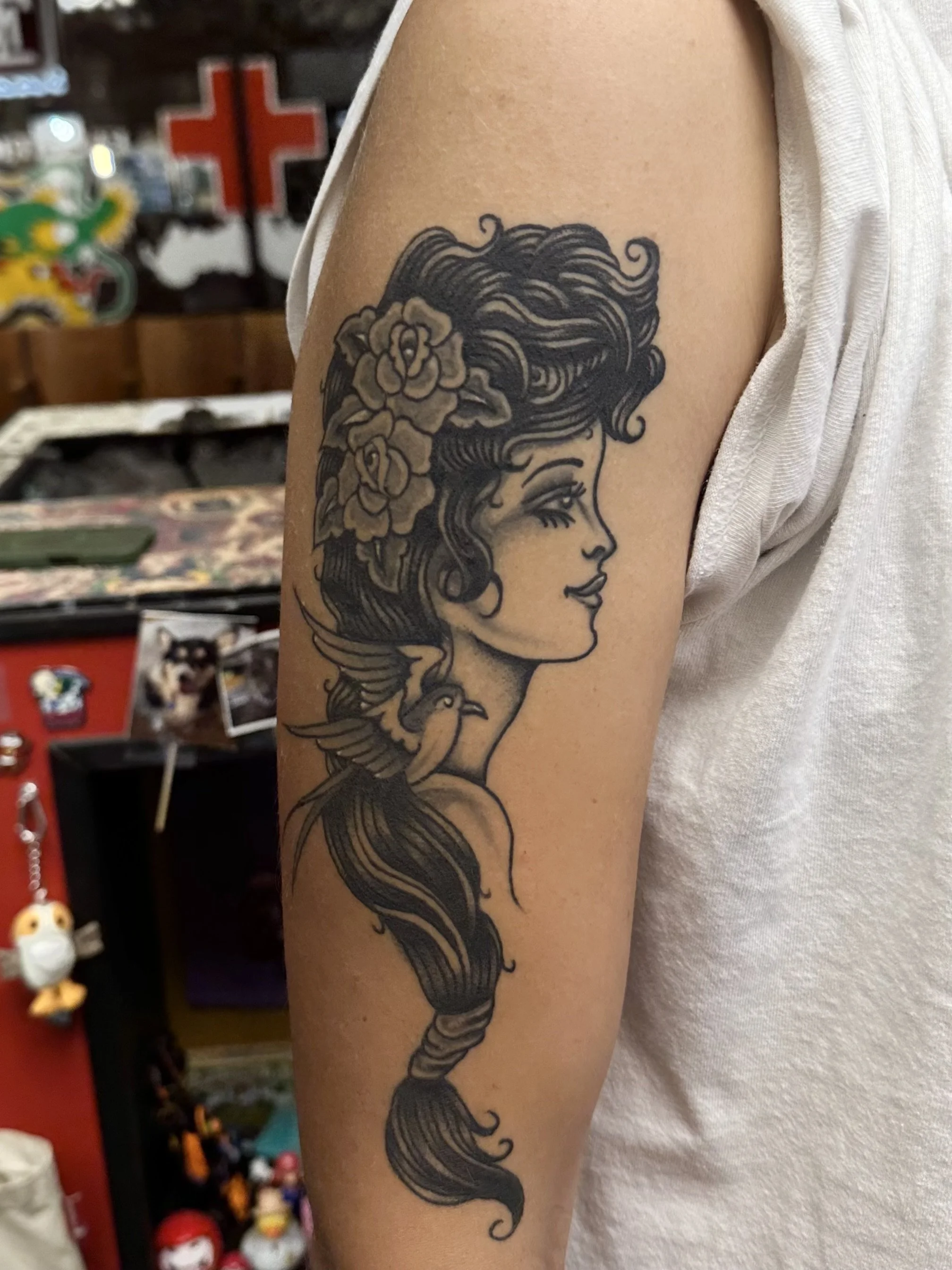 Tattoo of a woman's profile with flowing hair and flowers, with a bird at the bottom of the design, on a person's upper arm.
