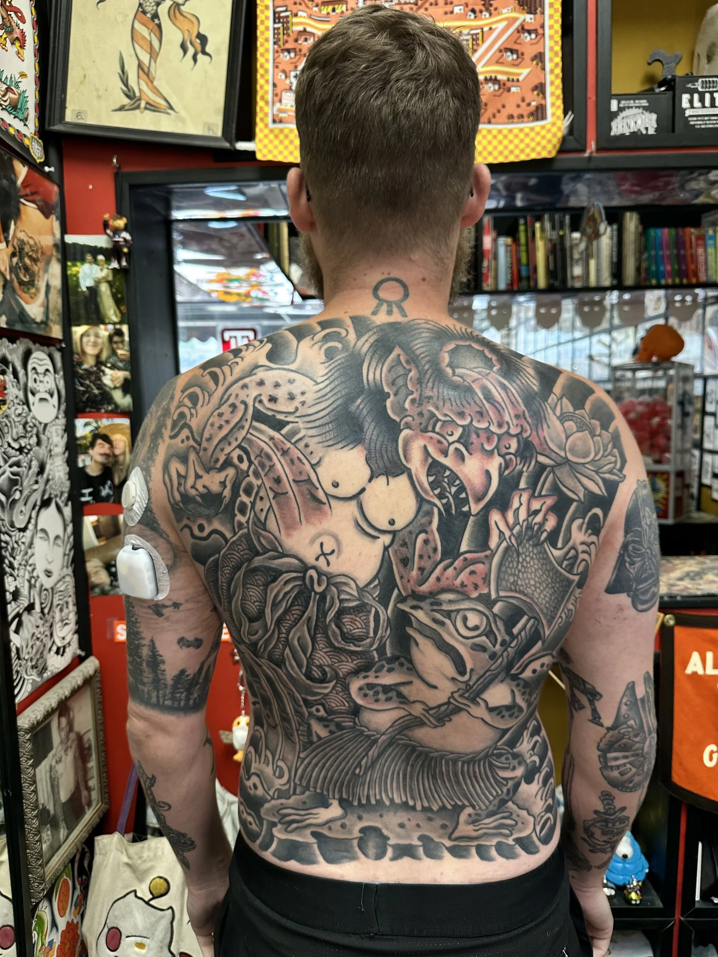 A shirtless man with tattoos covering his back and arms, standing in front of shelves filled with artwork and books in a store.