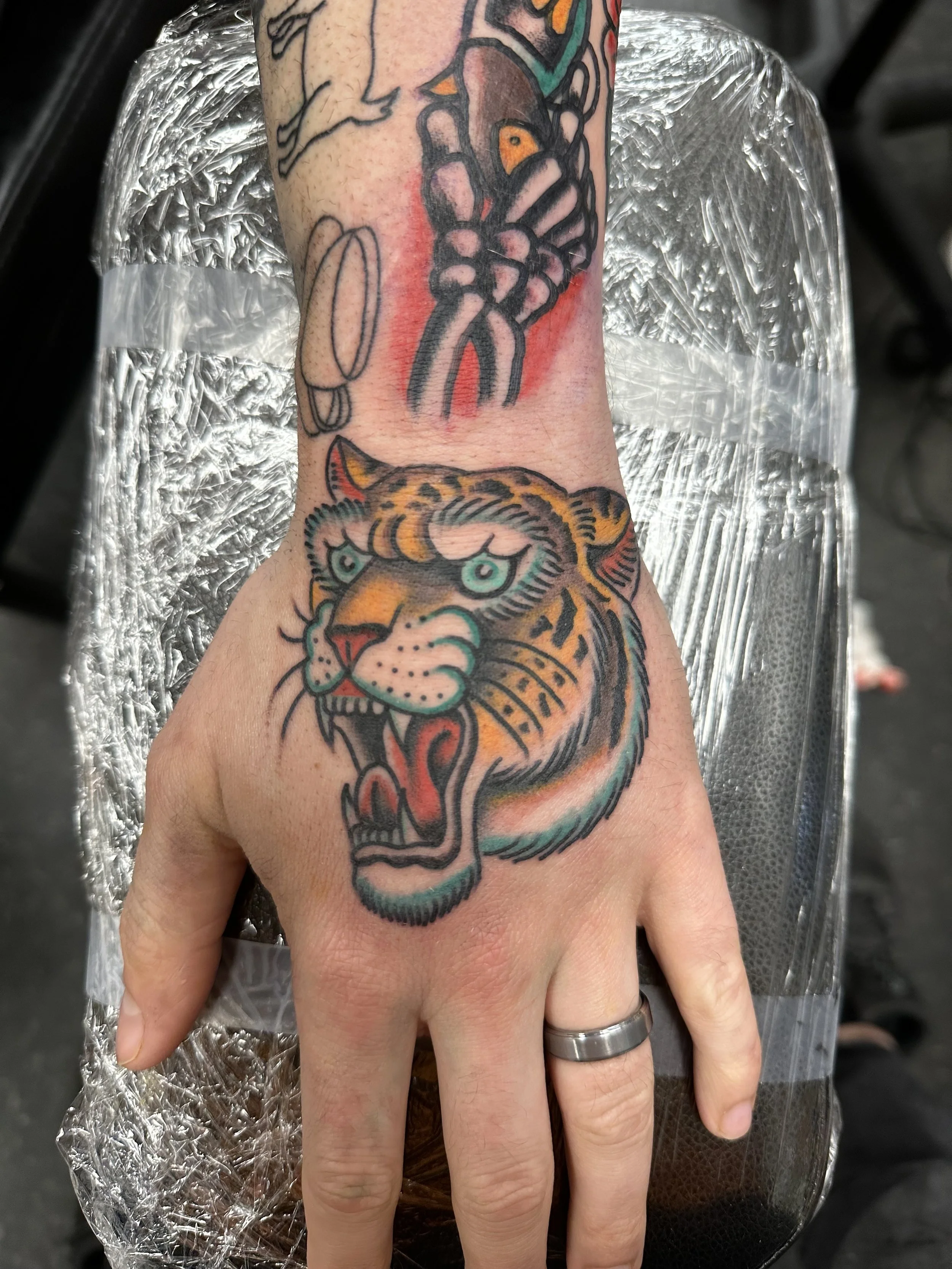 Tattoo of a tiger's head on a person's arm, with the tiger's mouth open showing teeth and tongue, and a tattoo of a butterfly on the arm.