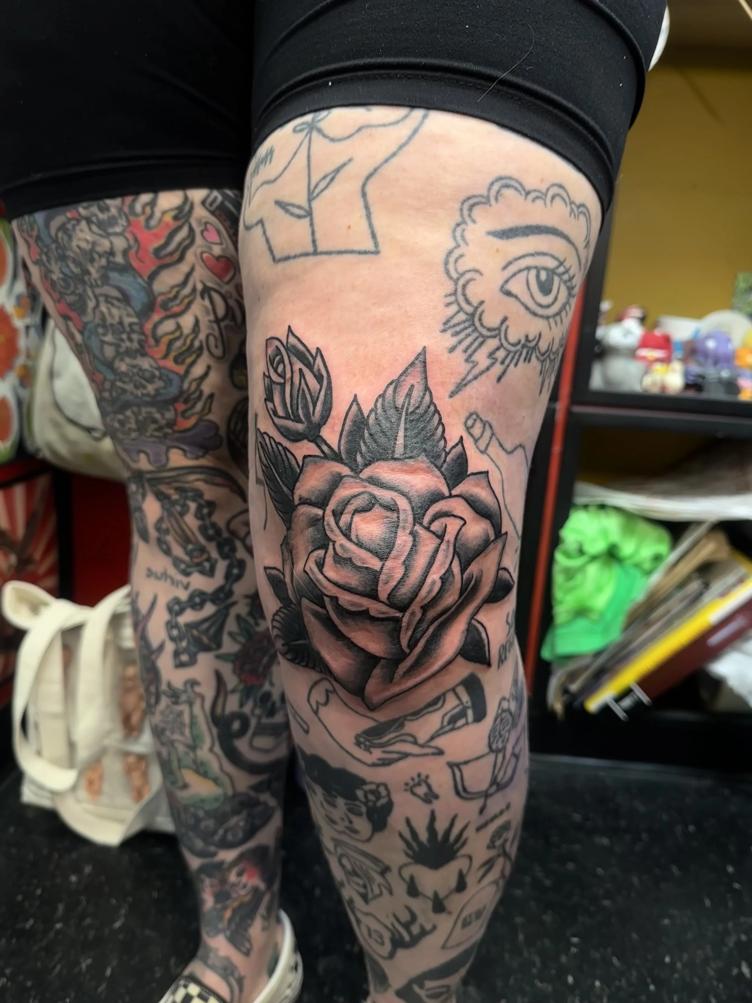 Close-up of a person's leg covered with various black and gray tattoos, including a large rose, eye with clouds, and other designs, with a background of storage shelves and various items.