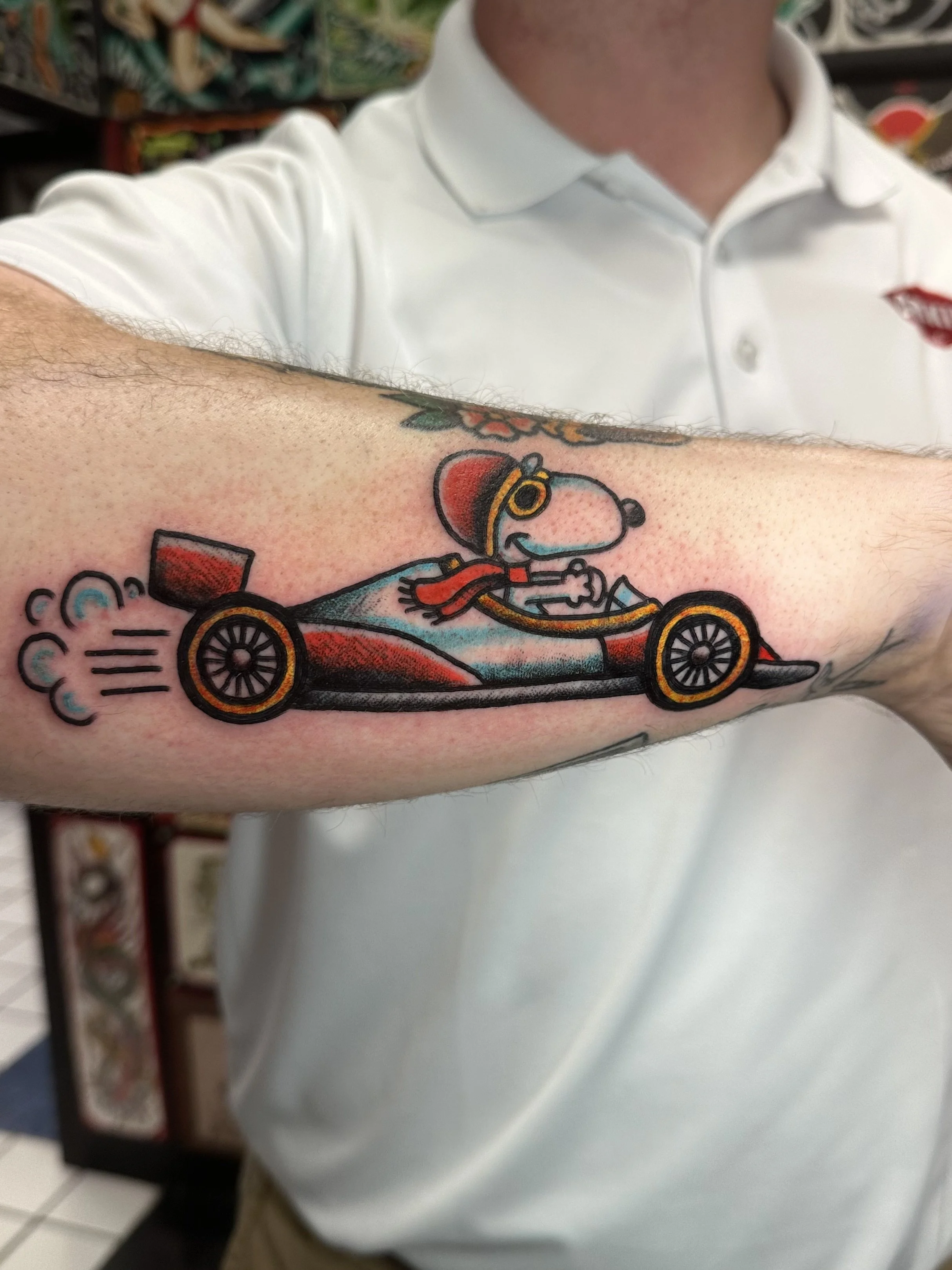 A person's arm with a cartoon tattoo of a racing dog in a red and black race car, wearing a red cap and scarf, with smoke or speed lines near the wheels.