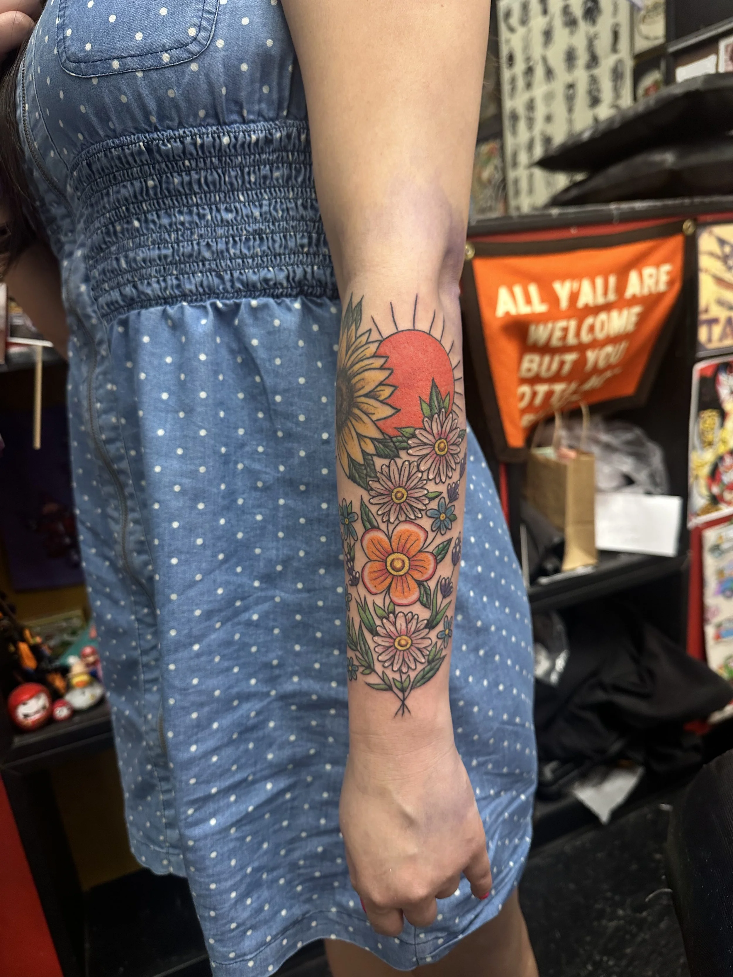 A person's arm with a colorful floral tattoo featuring sunflowers, pink, and orange flowers, extending from the wrist to the elbow, is visible. They are wearing a blue dress with white polka dots and smocking details on the back. In the background, a