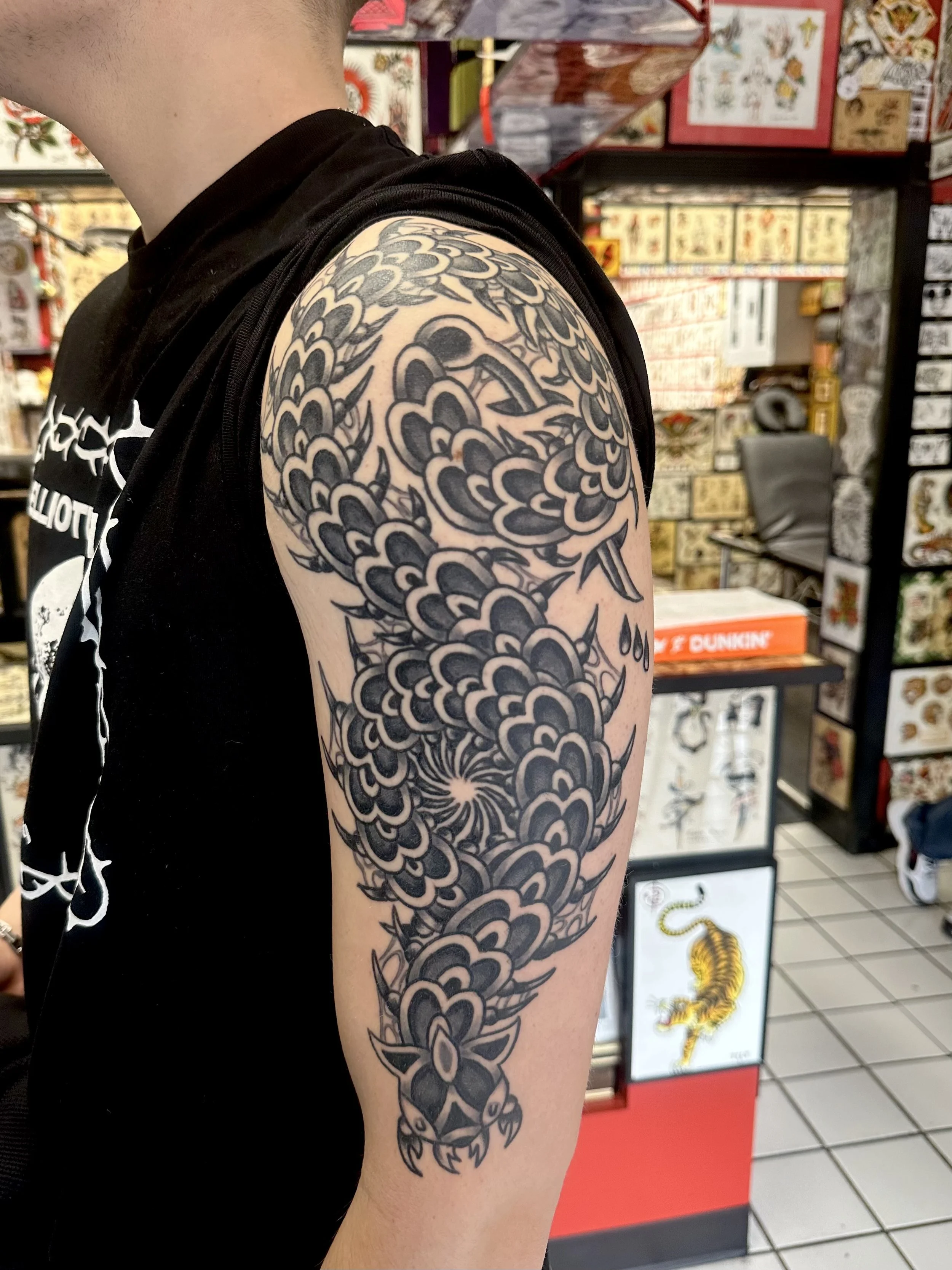 Person with a large dragon tattoo on their upper arm, sleeve style, in a tattoo shop with tattoo flash art on the walls.