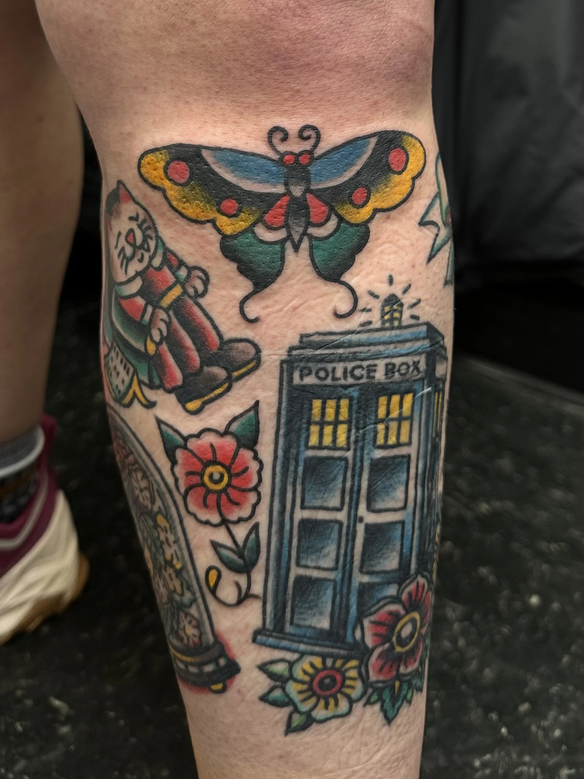 Colorful tattoo of a butterfly above a police box, with flowers and stars surrounding it.
