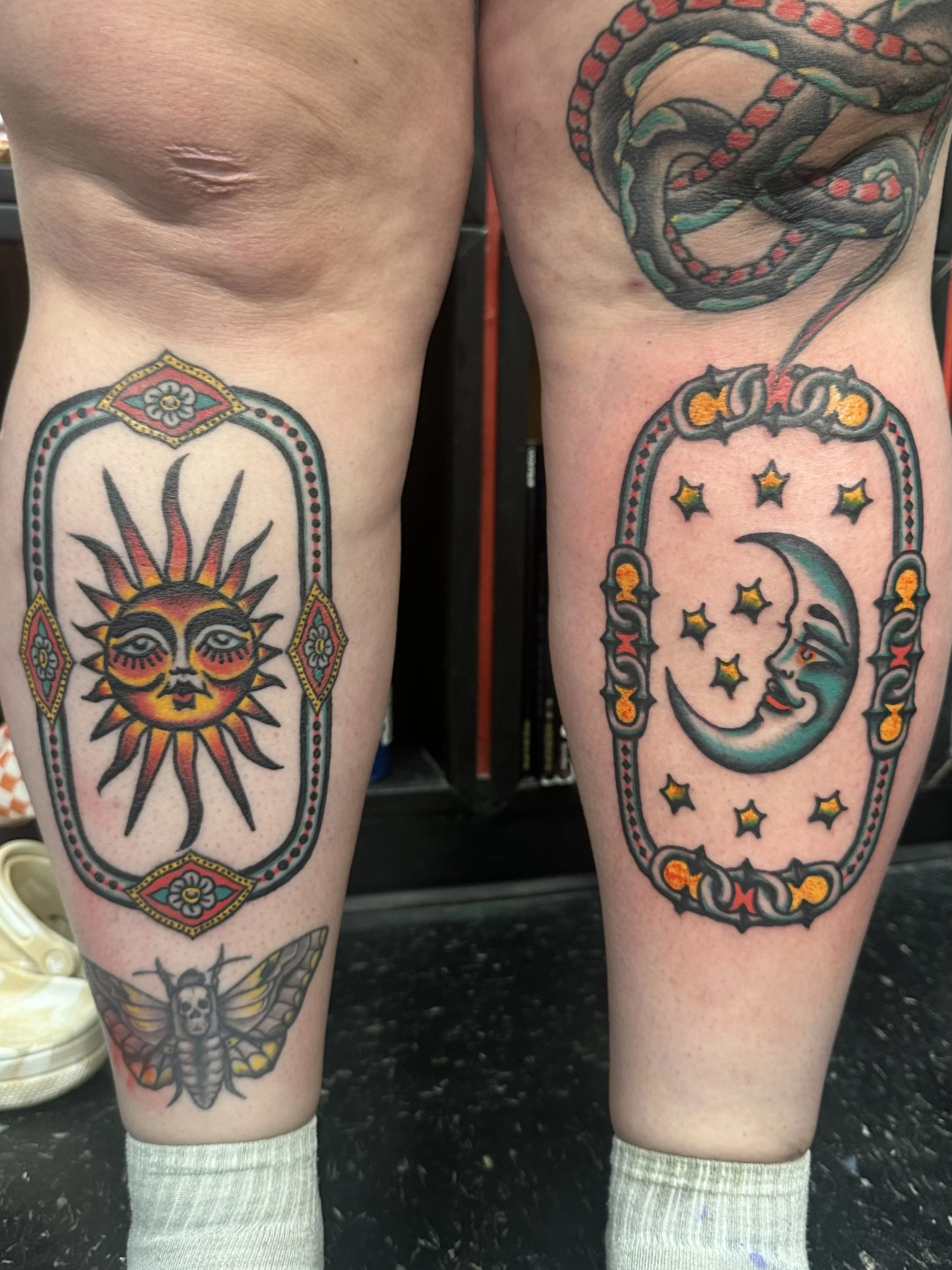 Close-up of two tattooed legs with colorful tattoos, including a sun face on the left thigh and a moon with stars on the right thigh.