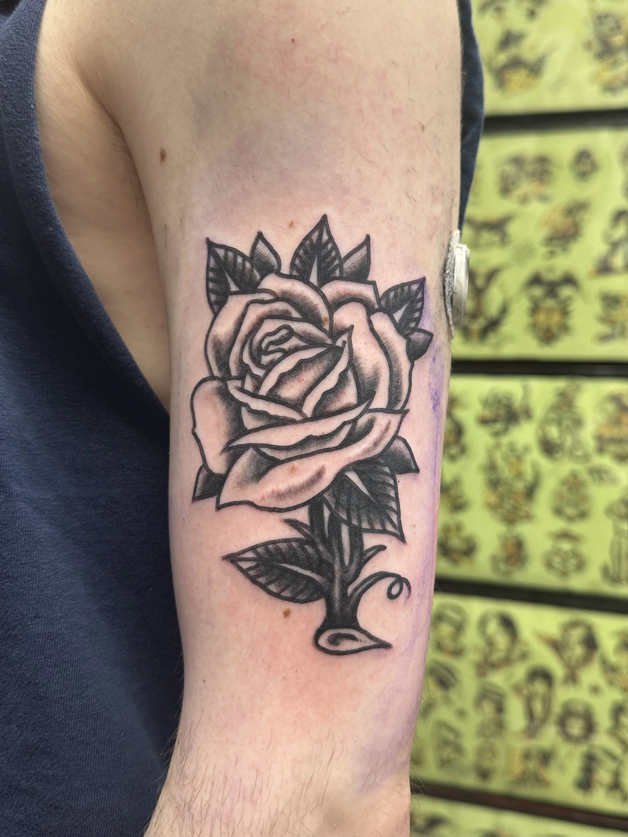 A black line tattoo of a rose with detailed petals and leaves on a person's upper arm.