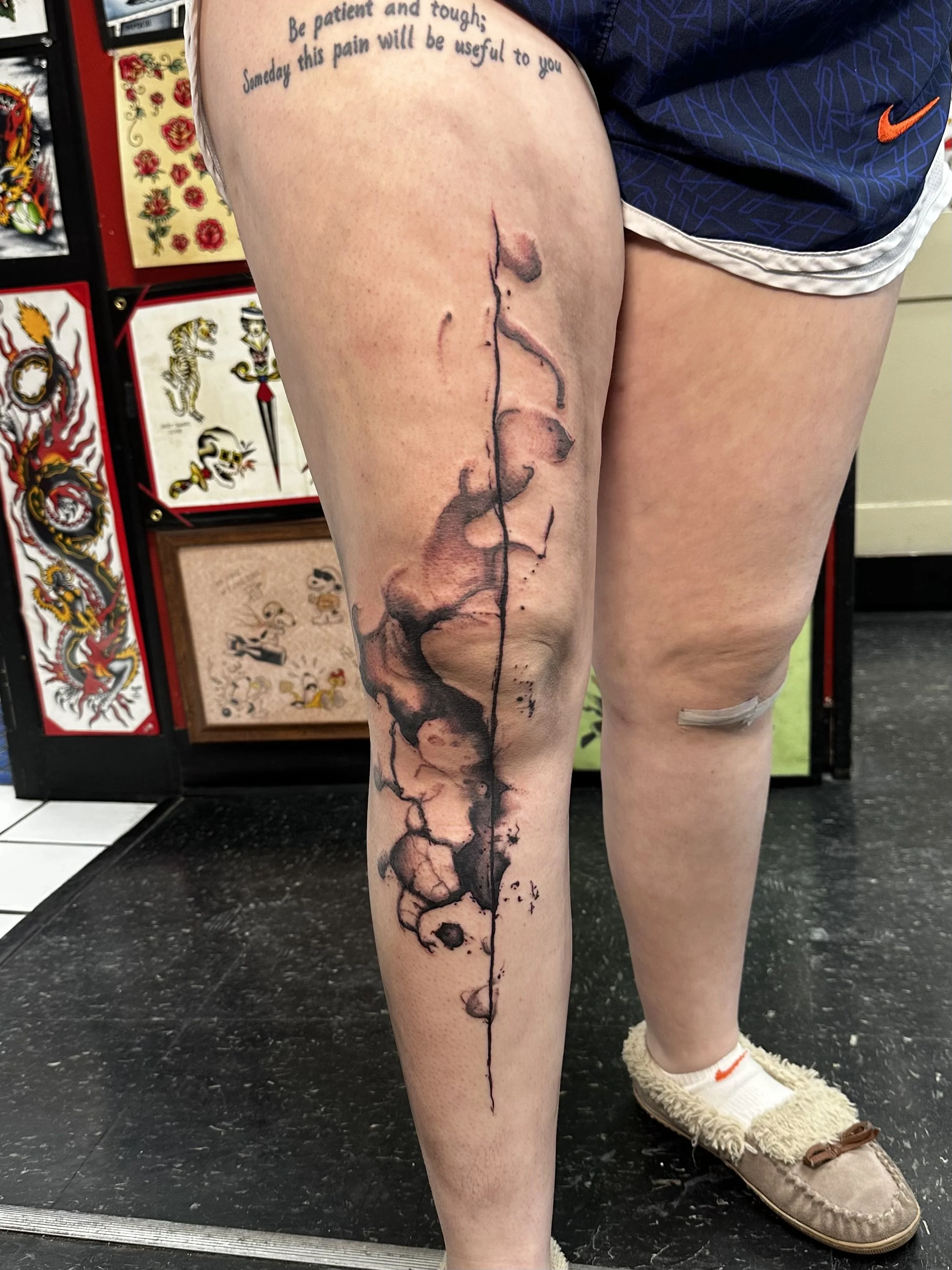 A person with a tattoo of a black ink inkpeaking in a continuous line from the thigh to the ankle, resembling a burning or melting effect. They are wearing beige slippers, white socks with orange stripes, and dark blue shorts. Tattoos and tattoo art 