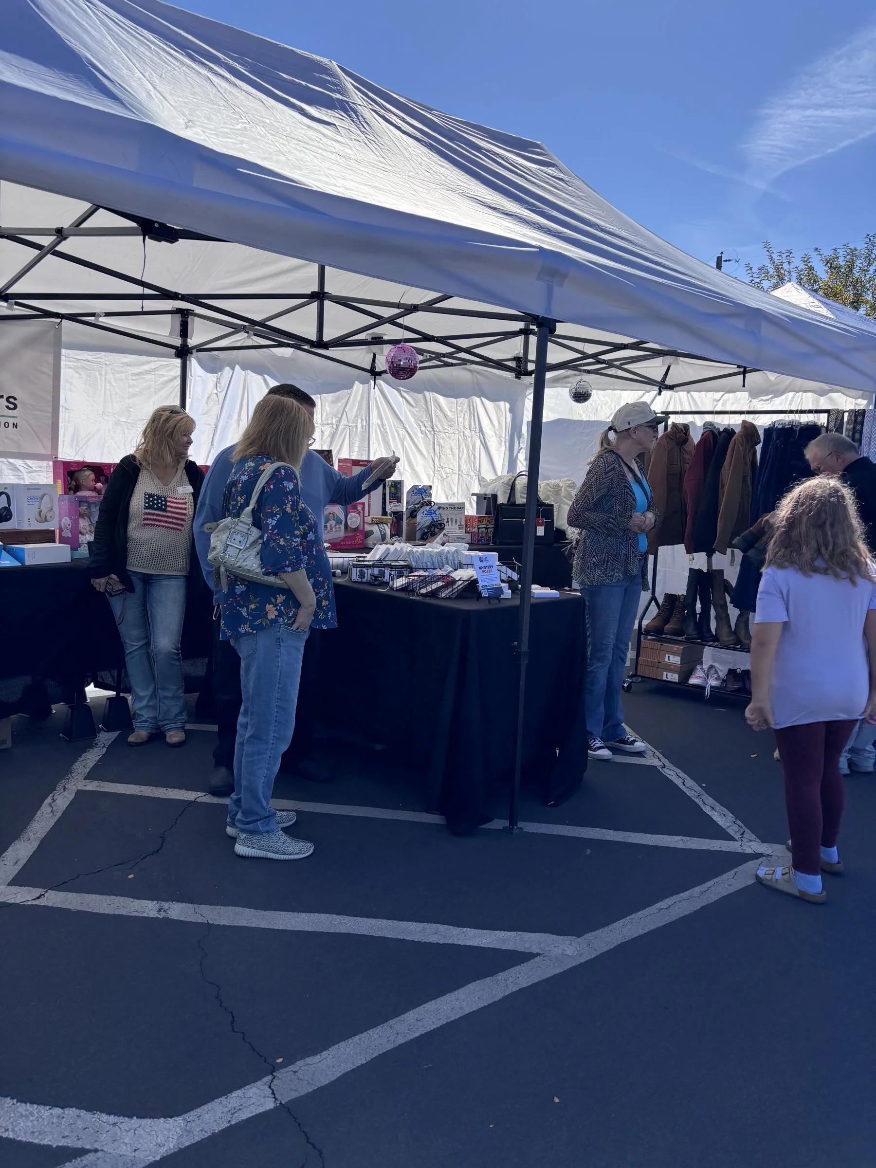 People shopping at a marketplace AEC Liquidators stall under a white tent on a sunny day with a clear blue sky, and some trees in the background.