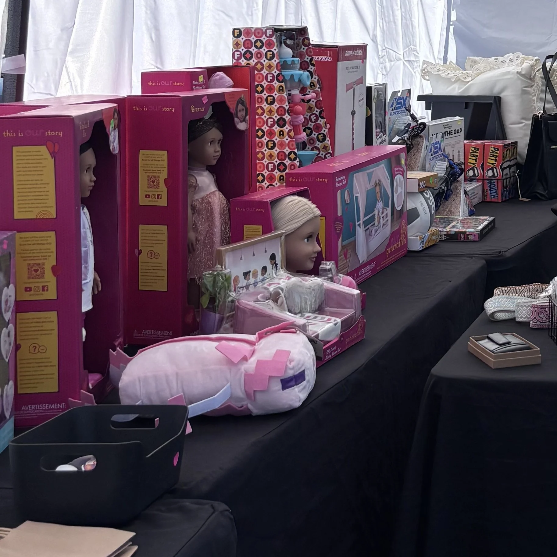 AEC Liquidators Overstock Display table with various boxed dolls and toys, including a doll with a pink dress and other boxed toys, in front of a white curtain backdrop.