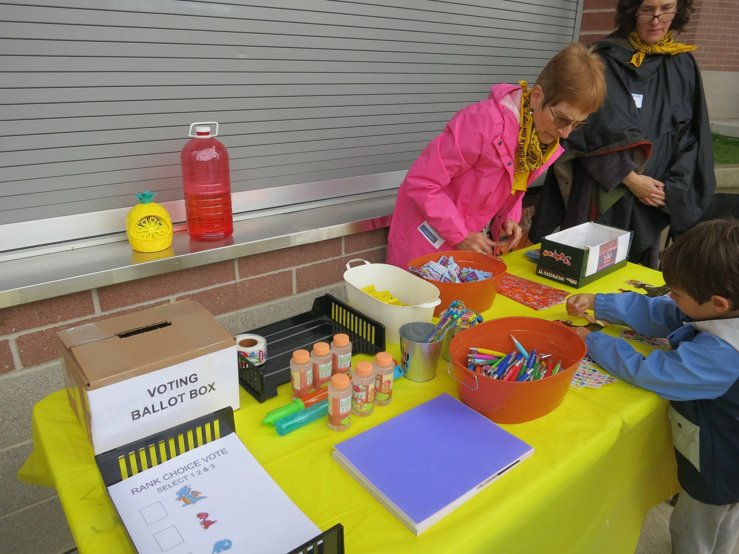 People at a voting event with a ballot box, various supplies, and signs on a yellow table.