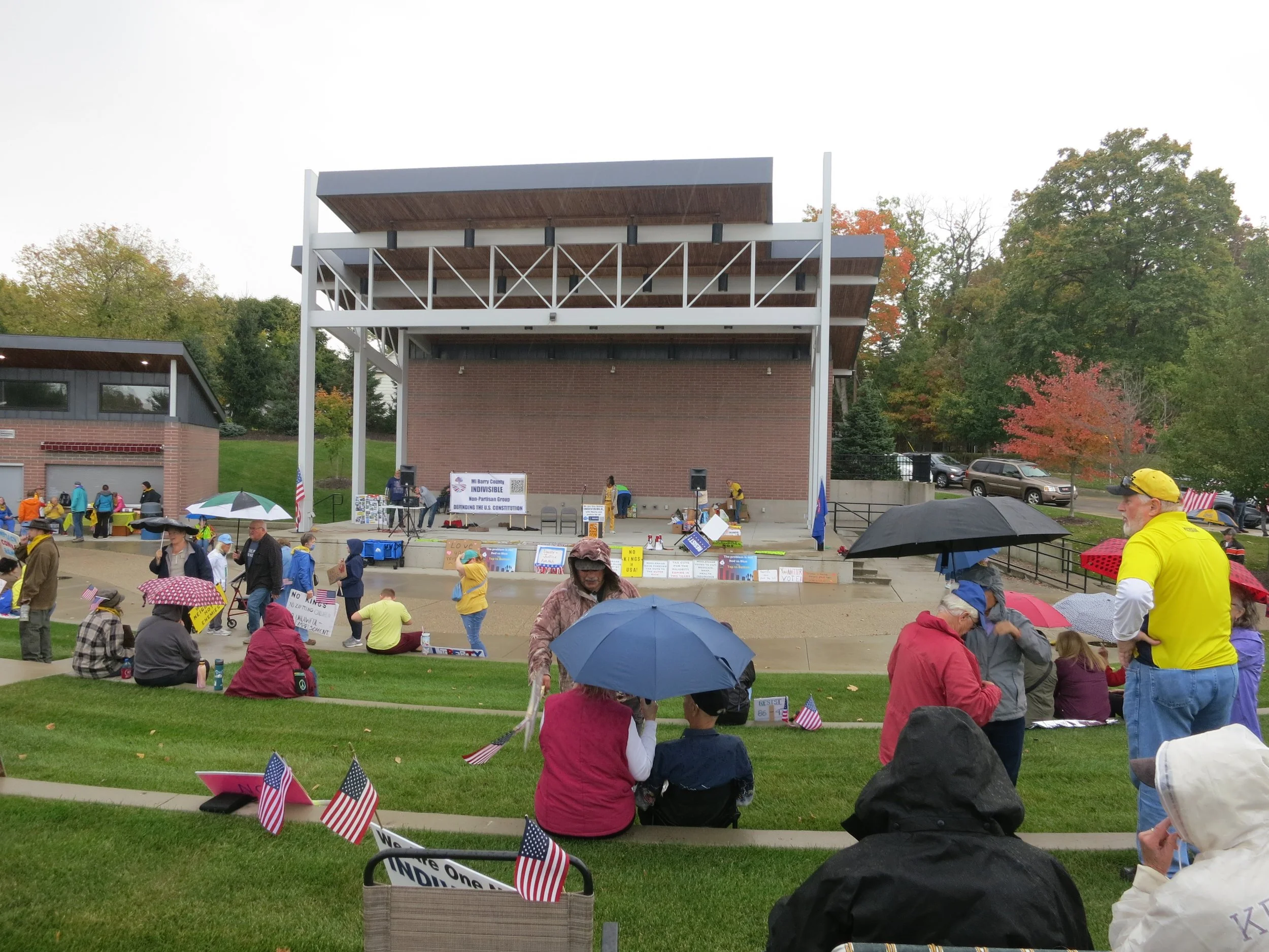 A crowd of people gathered on a lawn during a rainy day, holding umbrellas, with many small American flags and signs, with a stage and trees in the background.