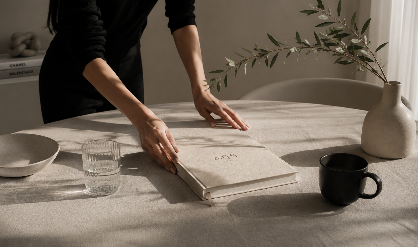 Person opening a beige notebook on a beige table, with a black mug, a glass of water, a white bowl, a beige vase with a branch, and some books in the background.