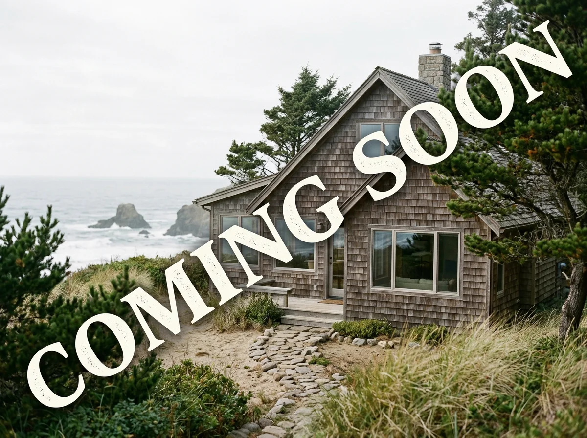 A seaside house with a wooden exterior, surrounded by trees and bushes, overlooking the ocean with rocky formations in the background, and a stone pathway leading to the entrance.