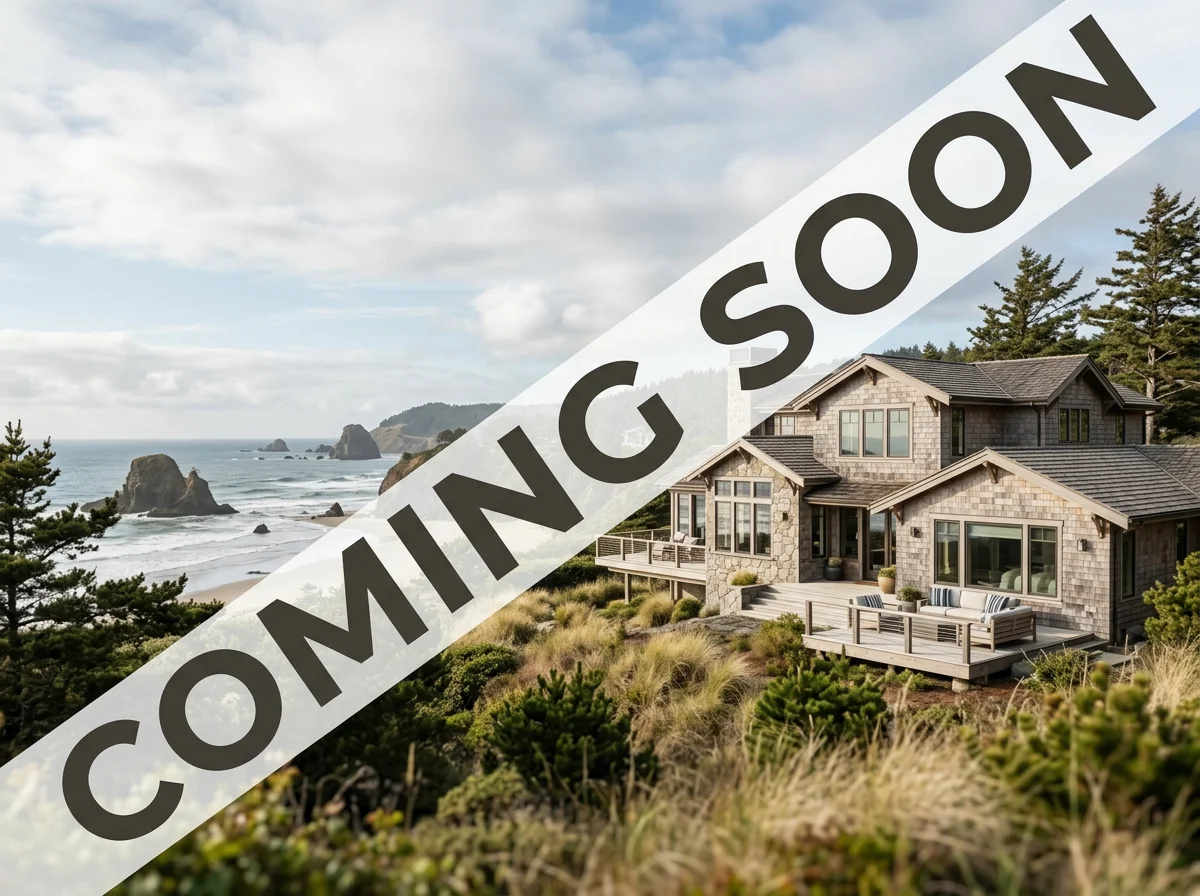 A large two-story coastal house with a deck, surrounded by bushes and trees, overlooking a beach with rocky formations and the ocean.
