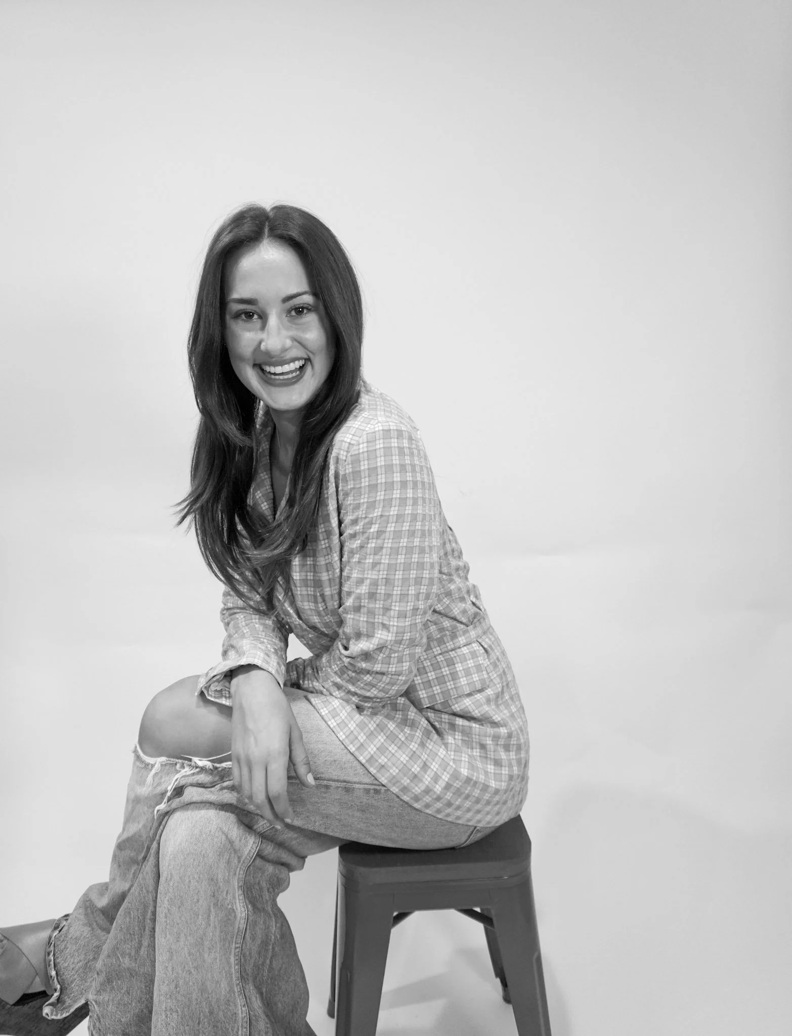 Black and white photo of a young woman with long hair, smiling, sitting on a stool against a plain background, wearing a checkered shirt and ripped jeans.