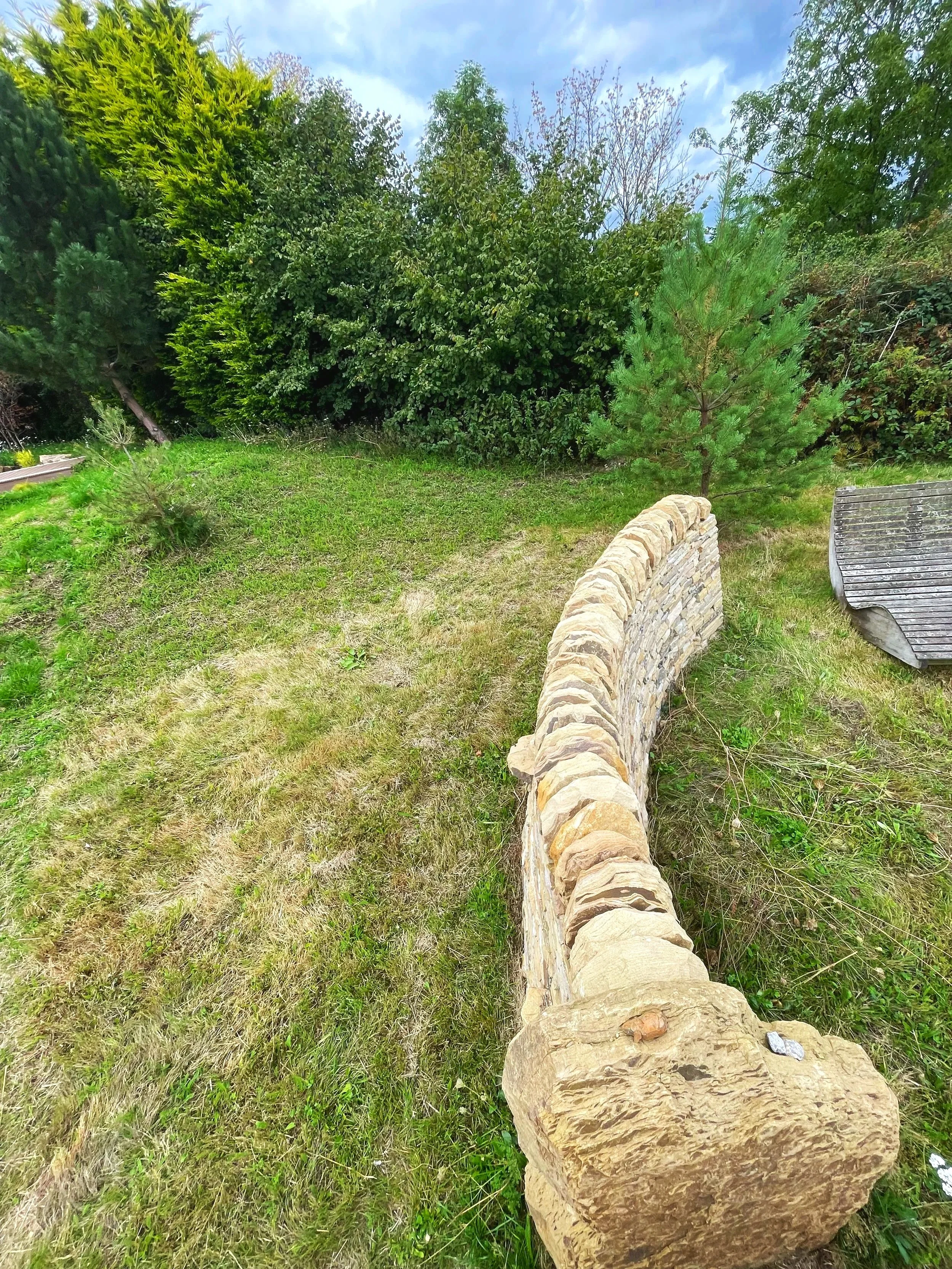 A large log lying on a grassy patch of land with a stone wall at its side, surrounded by green bushes and trees under a partly cloudy sky.