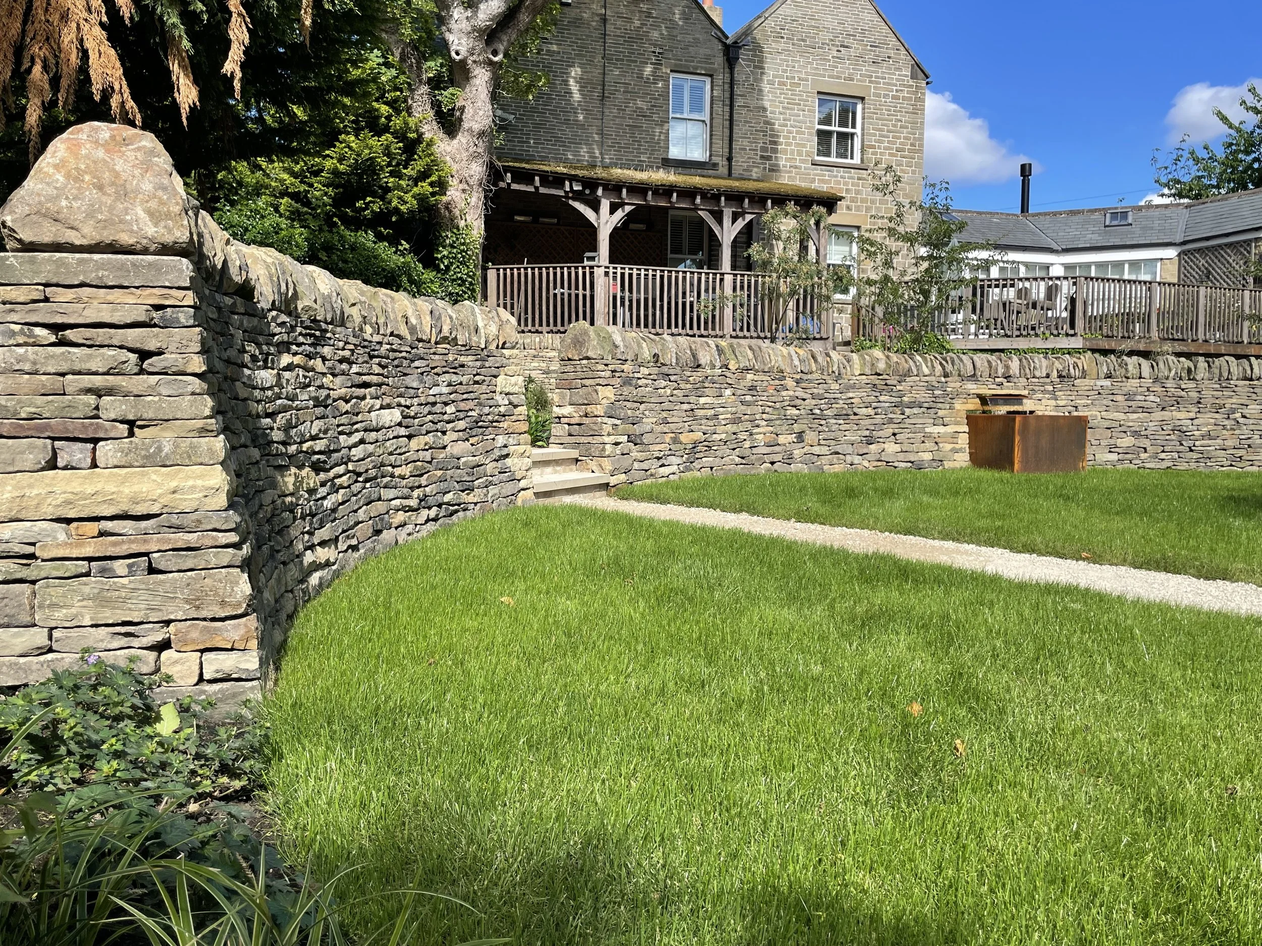 A backyard with a lush green lawn, a stone wall, and a wooden deck attached to a brick house. The house has two stories with multiple white-framed windows and a covered porch area. There are trees and bushes around the yard, and a blue sky with a few