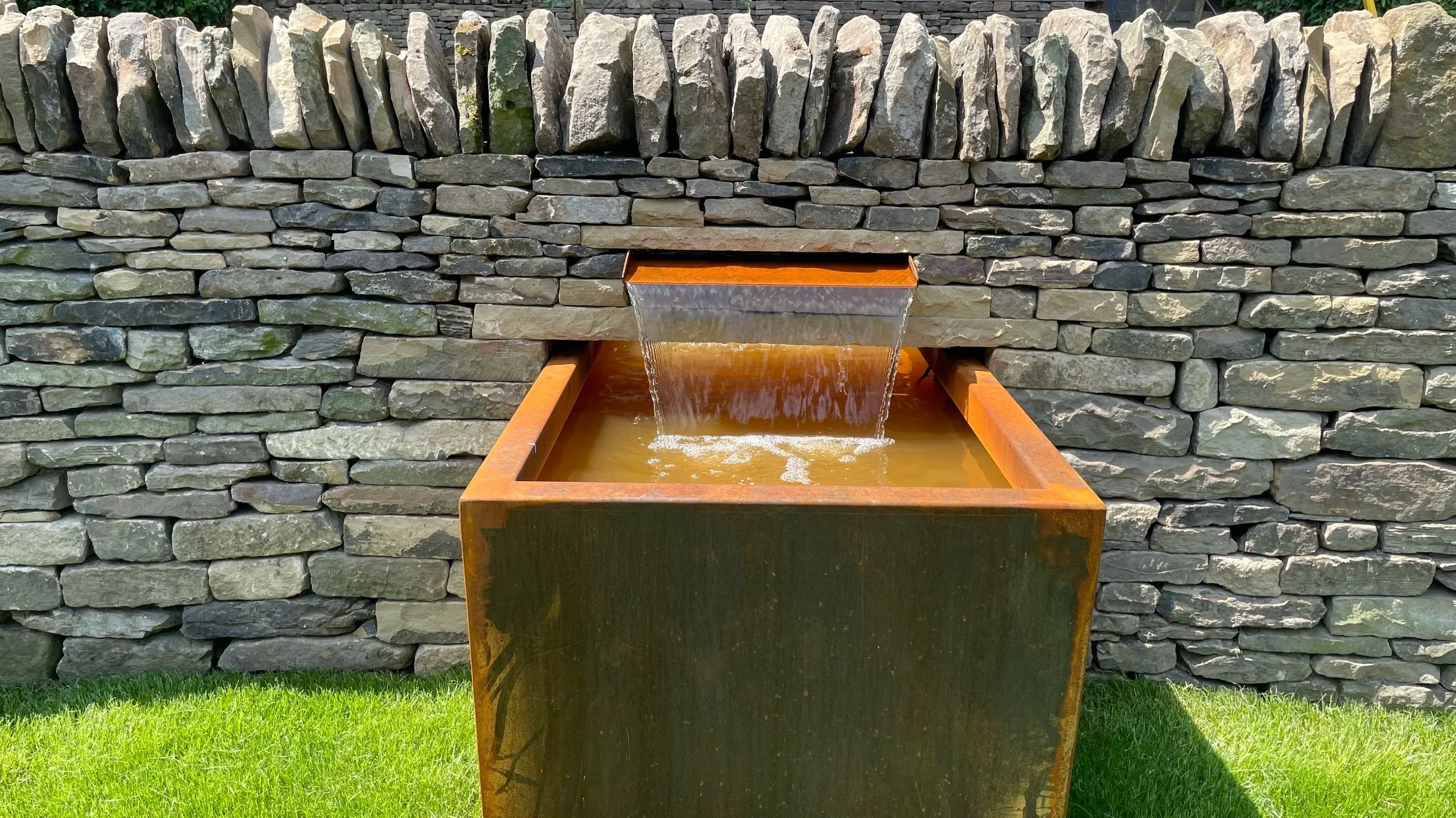 A decorative water feature made of dark stone stones with water flowing from a rectangular metal spout into a rust-colored metal basin, set against a tall stacked stone wall in a garden.