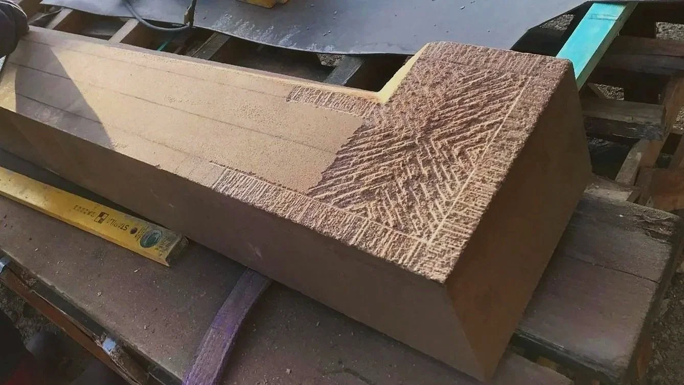 A woodworking project in progress showing a large rectangular block of wood with a carved pattern on one end, resting on a workbench with measuring tools nearby.