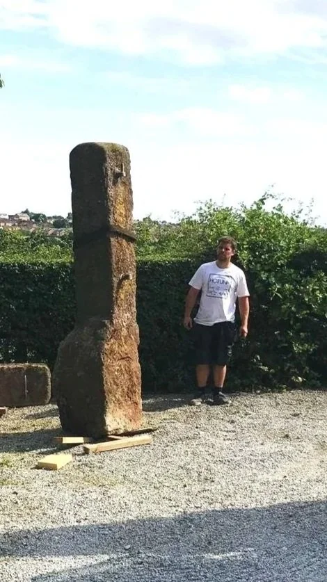 A person standing next to an abstract stone sculpture consisting of two large, irregularly shaped stones balanced vertically on a gravel ground with greenery and a cloudy sky in the background.
