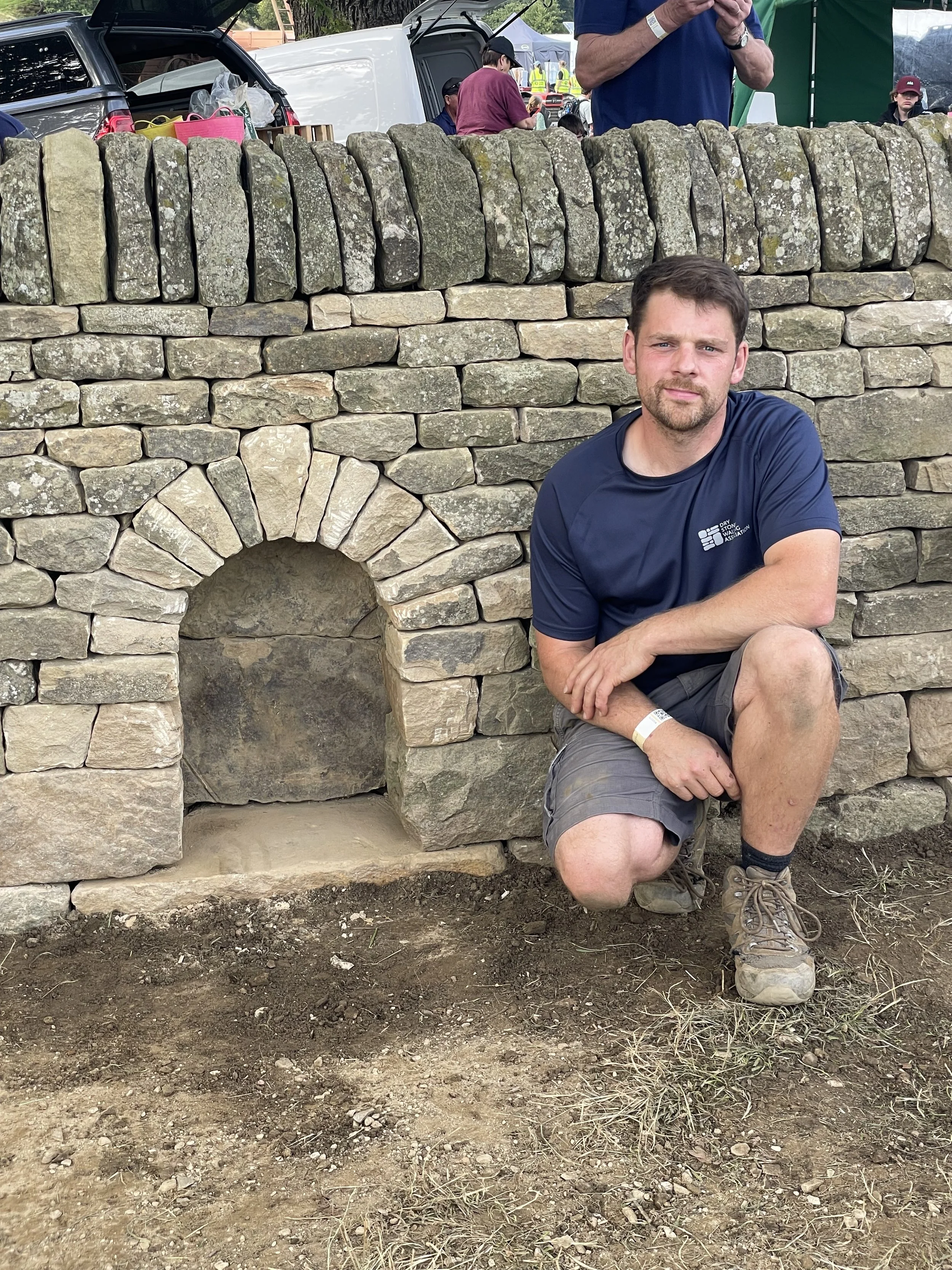 A man with short brown hair and a beard, wearing a dark blue t-shirt and gray shorts, is kneeling in front of a stone wall with an arch opening. The wall is made of gray and beige stones. The man is outdoors at what appears to be a festival or outdoor event, with people and vehicles visible in the background.