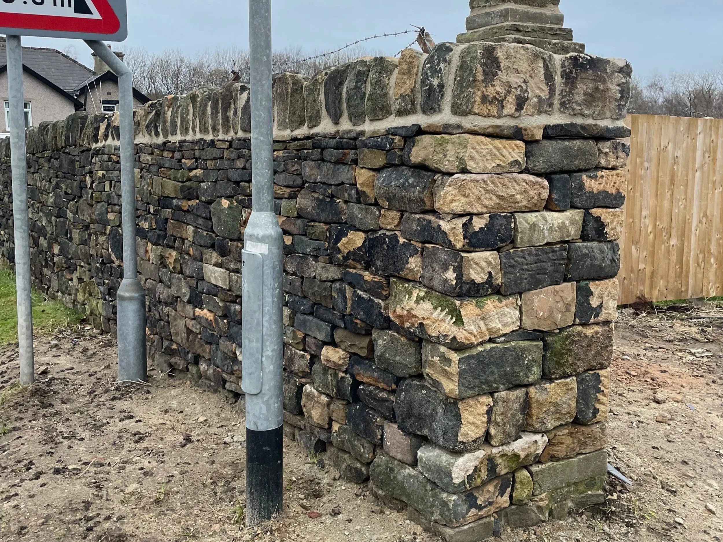 A stone and brick wall under construction with scaffolding poles in front, surrounding a residential area with houses and trees.