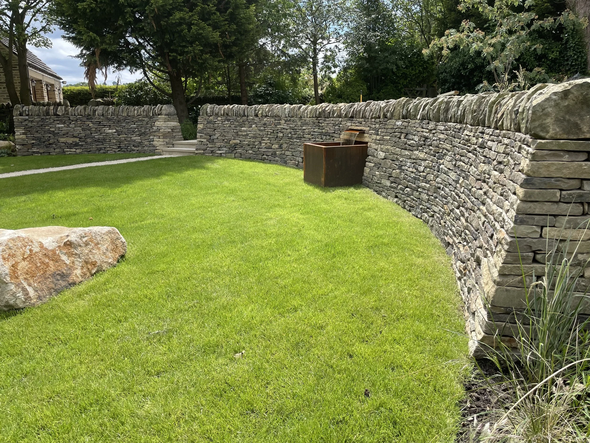 A stone retaining wall with steps, a rusty decorative fountain, green grass, trees, and a house in the background under cloudy sky.