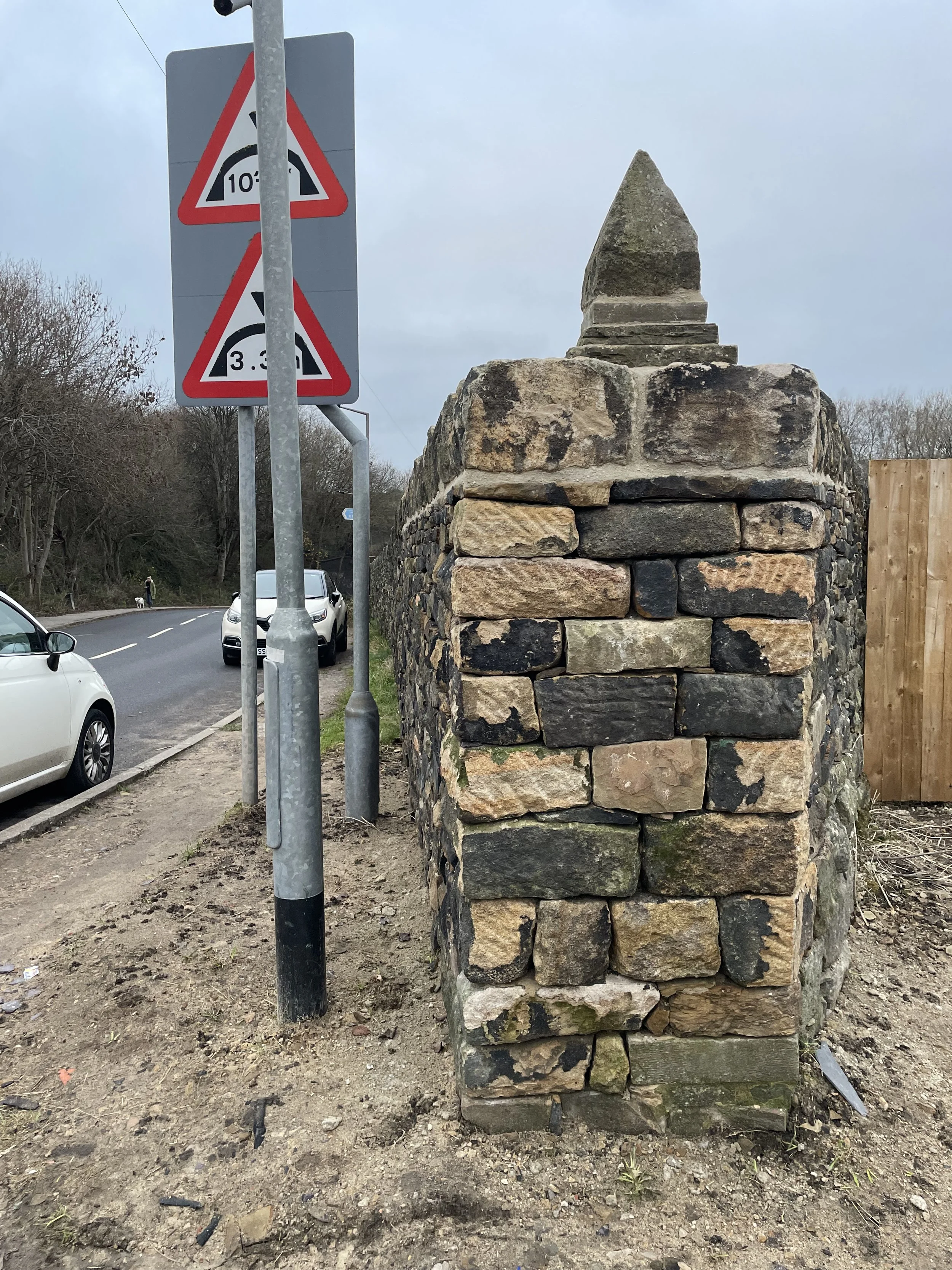 Street scene with a traffic sign and a stone brick wall, cars parked along the road, with someone walking a dog in the background on a cloudy day.