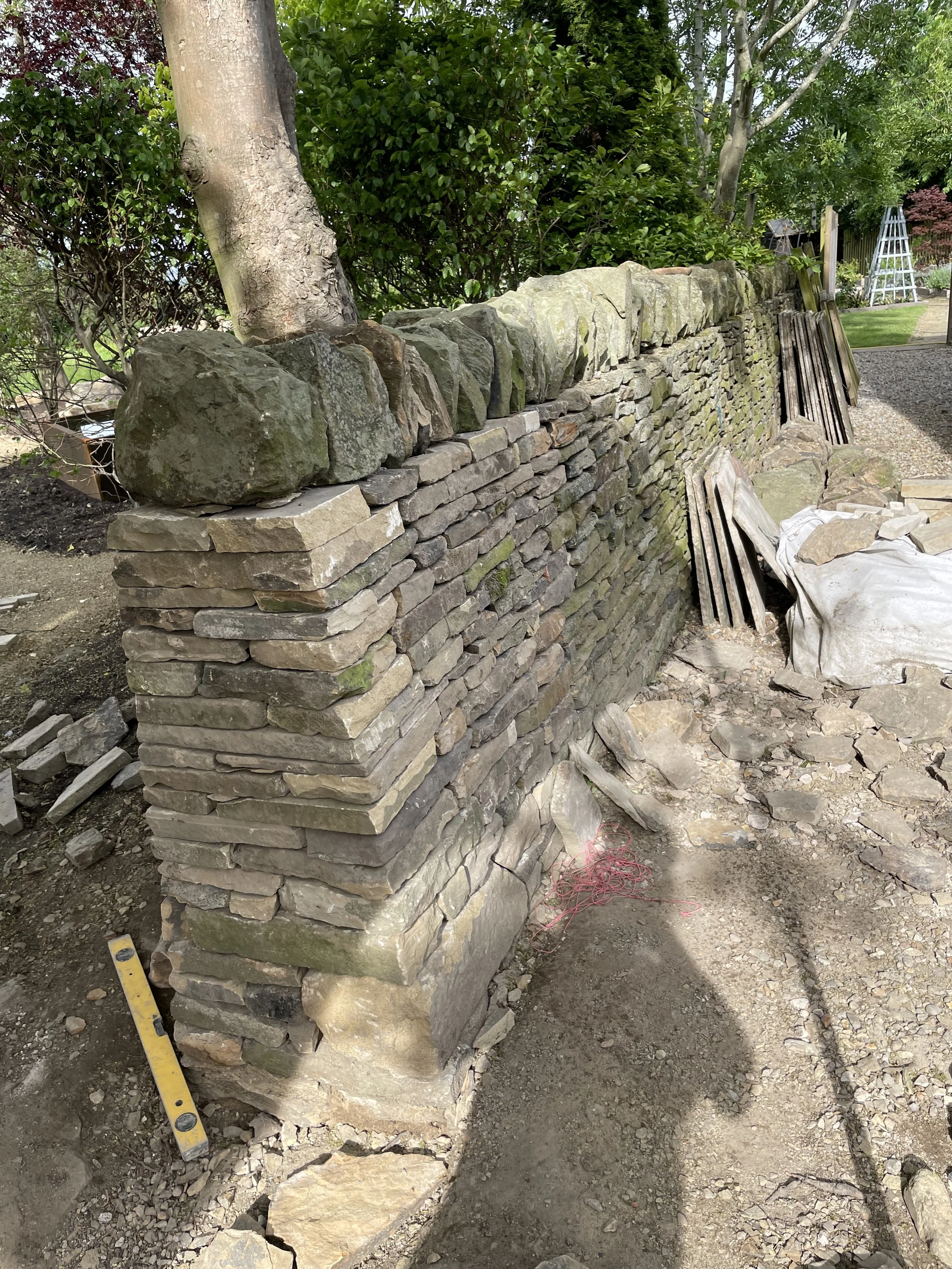 A stone wall under construction with large rocks on top of smaller, flat stones, in a garden setting with trees and green shrubbery in the background. Construction tools and materials are nearby.