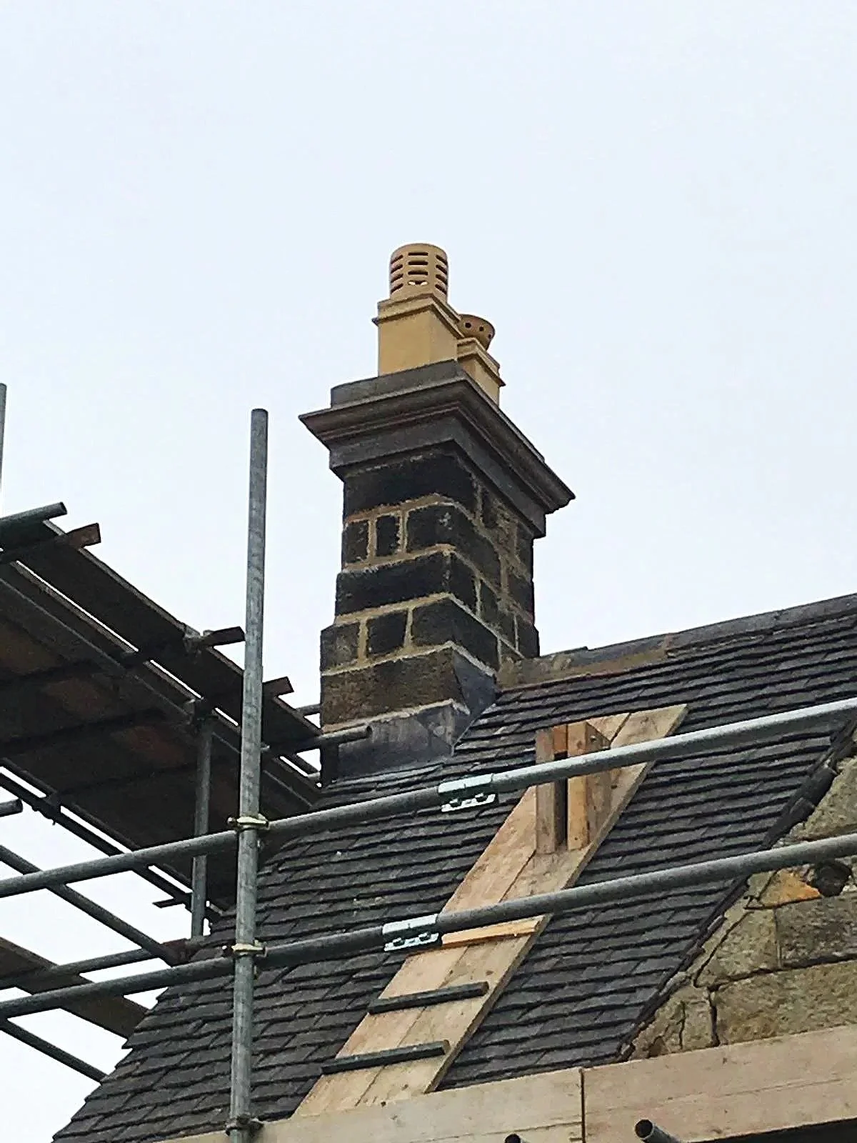 Chimney on a roof under construction with scaffolding and a wooden ladder, surrounded by building materials.