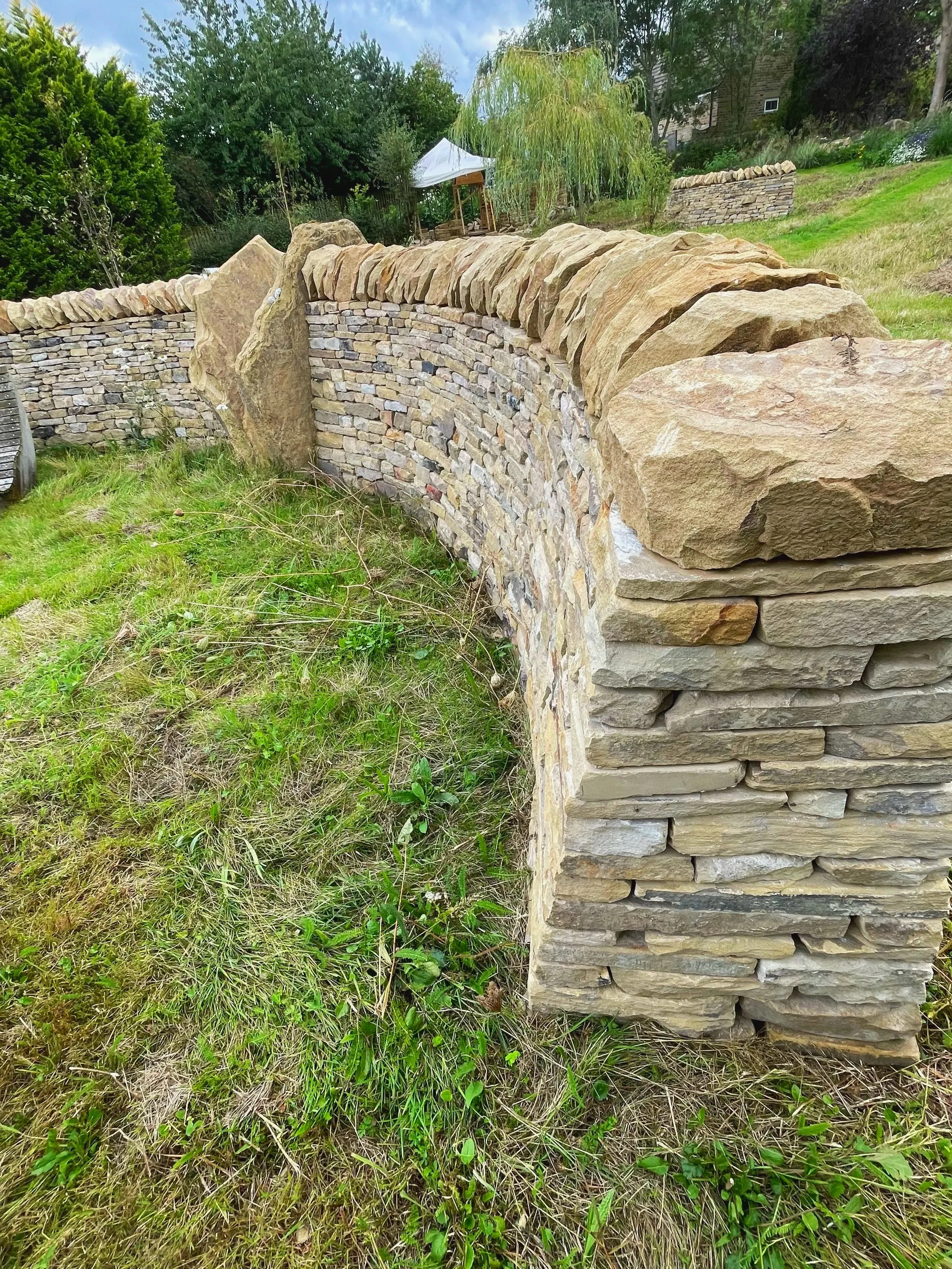 Stone wall with large natural rocks on top, green grass and small plants in foreground, trees and a small outdoor canopy in the background.