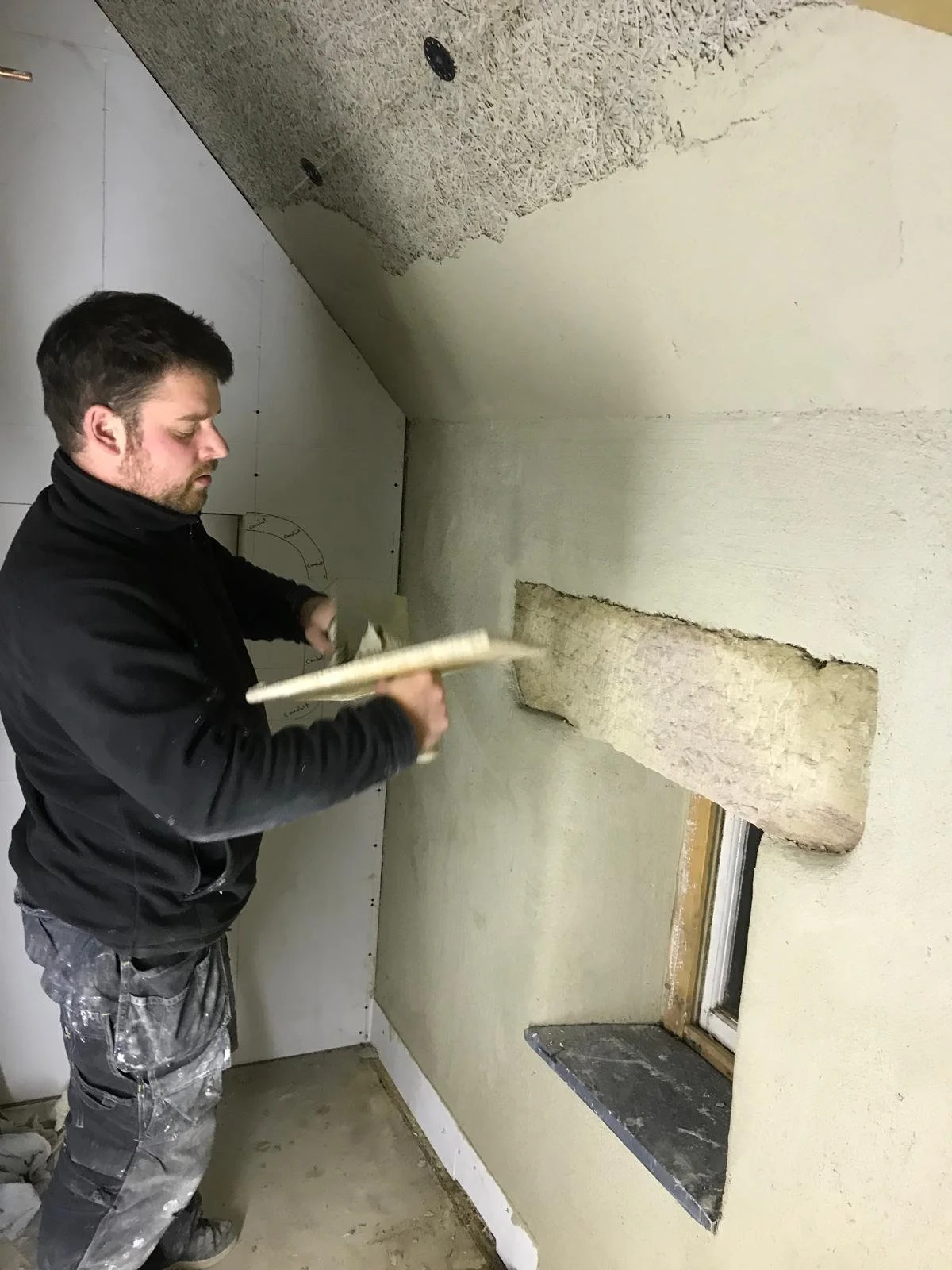 Man inspecting a wall with a large hole, holding a notebook, in a room under renovation.