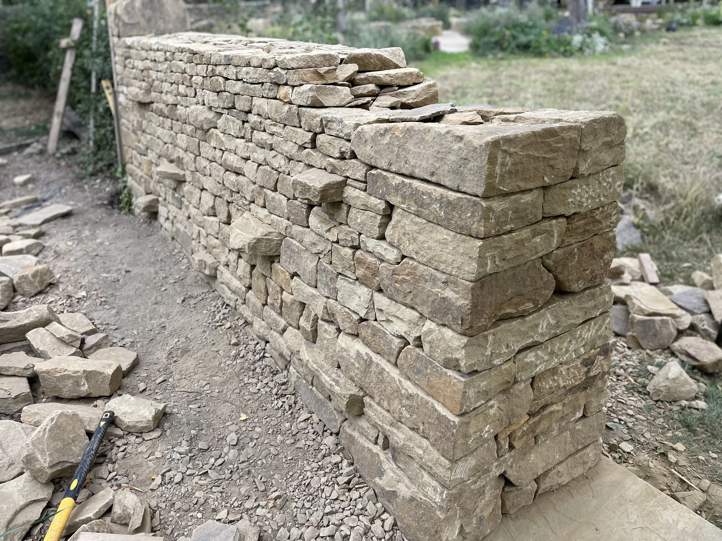 A partially constructed dry stone wall made of rectangular and irregularly shaped tan and beige stones in a garden or outdoor setting.