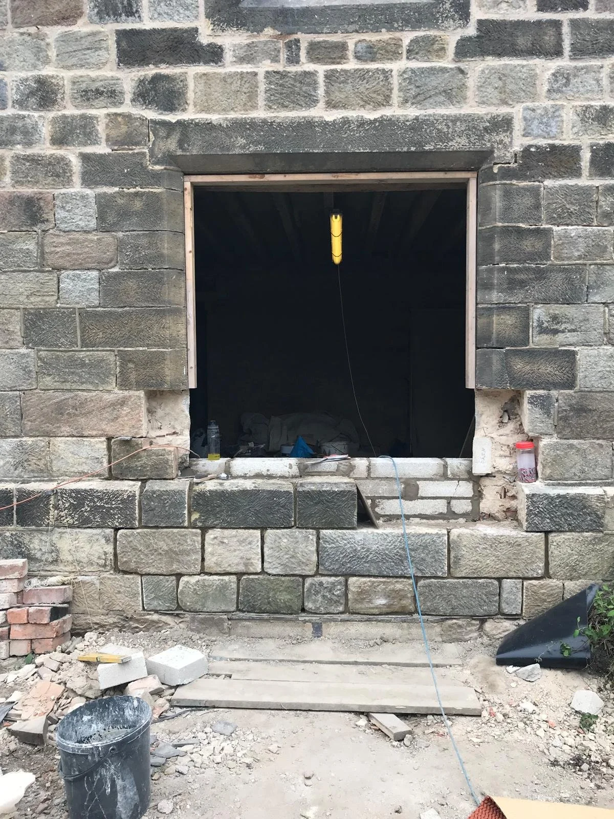 Construction site showing a partially built brick wall with an opening for a window. Construction tools, debris, and materials are scattered on the ground.