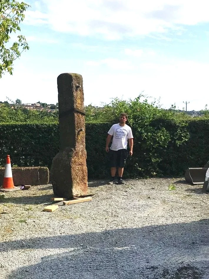A man standing outdoors next to a tall, vertical stone sculpture on a small wooden base, with a hedge and trees in the background.