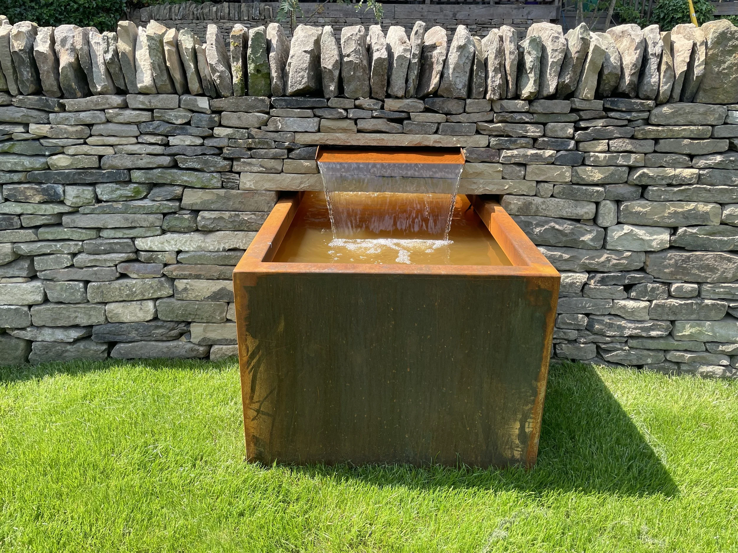 A stone wall with a water feature in front. The water flows from a rectangular opening into a rust-colored metal basin surrounded by green grass.