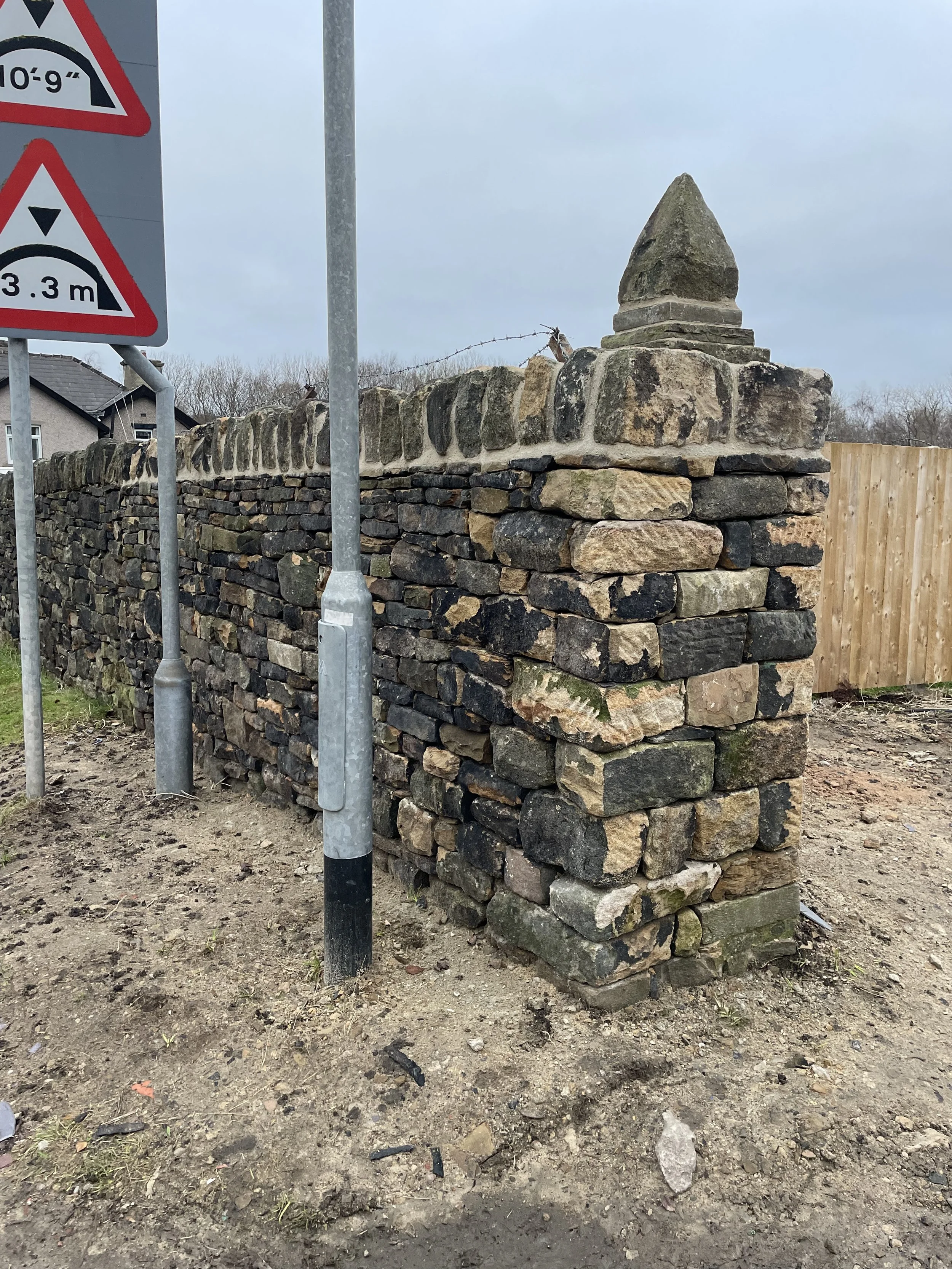 A stone wall with a decorative spire on top, situated outdoors next to a wooden fence and street signs, with a background of trees and overcast sky.