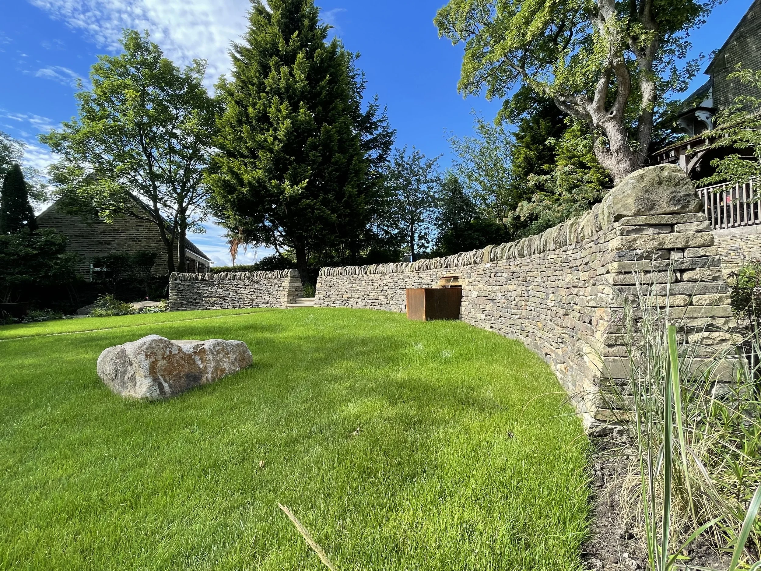 A landscaped yard with a lush green lawn, a large rock, a low stone wall, and tall trees with green foliage under a bright blue sky.