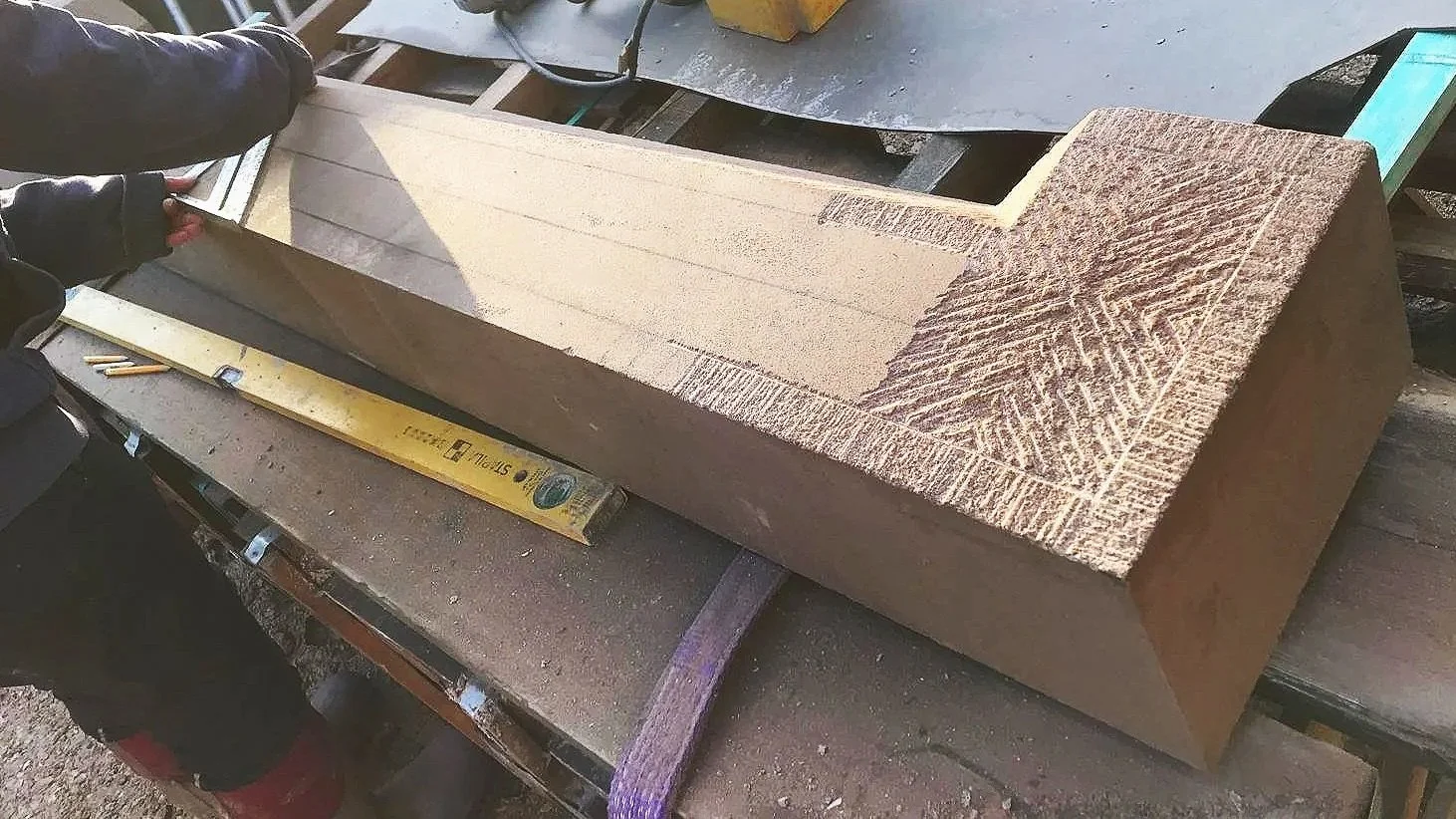 A person using a yellow level tool to measure a rectangular carved stone block with detailed patterns on its edges, placed on a workbench.