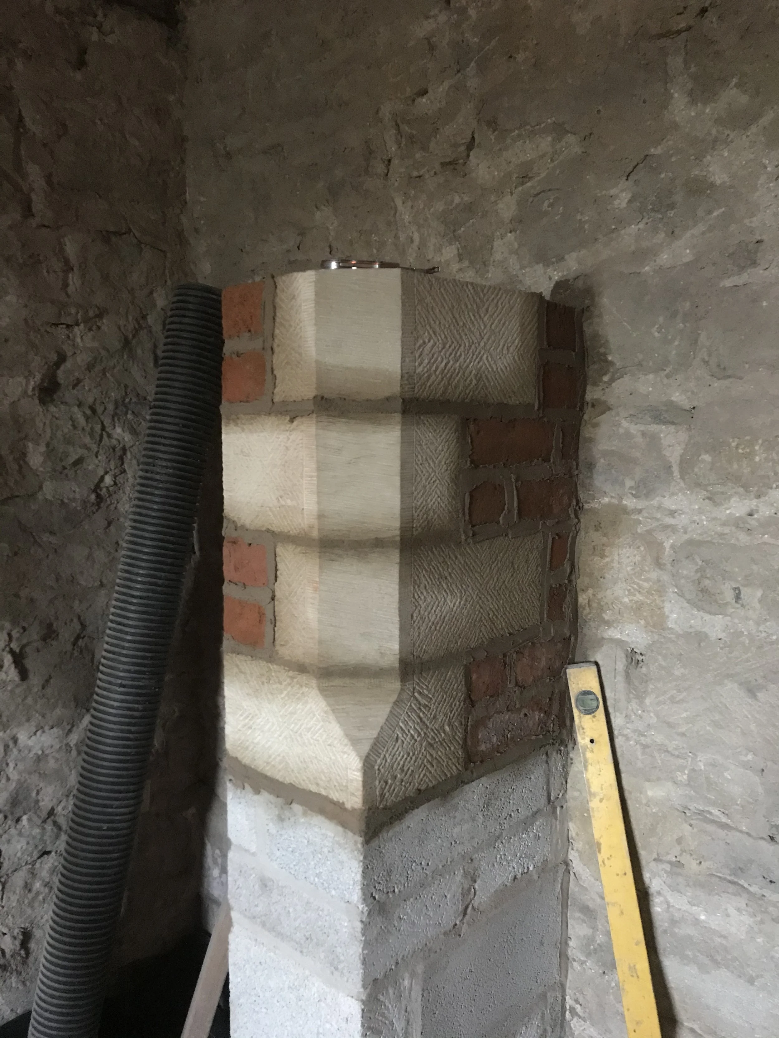 Construction site showing a concrete wall with bricks and a large black flexible pipe attached, a yellow level on the ground, and rough cement floor.
