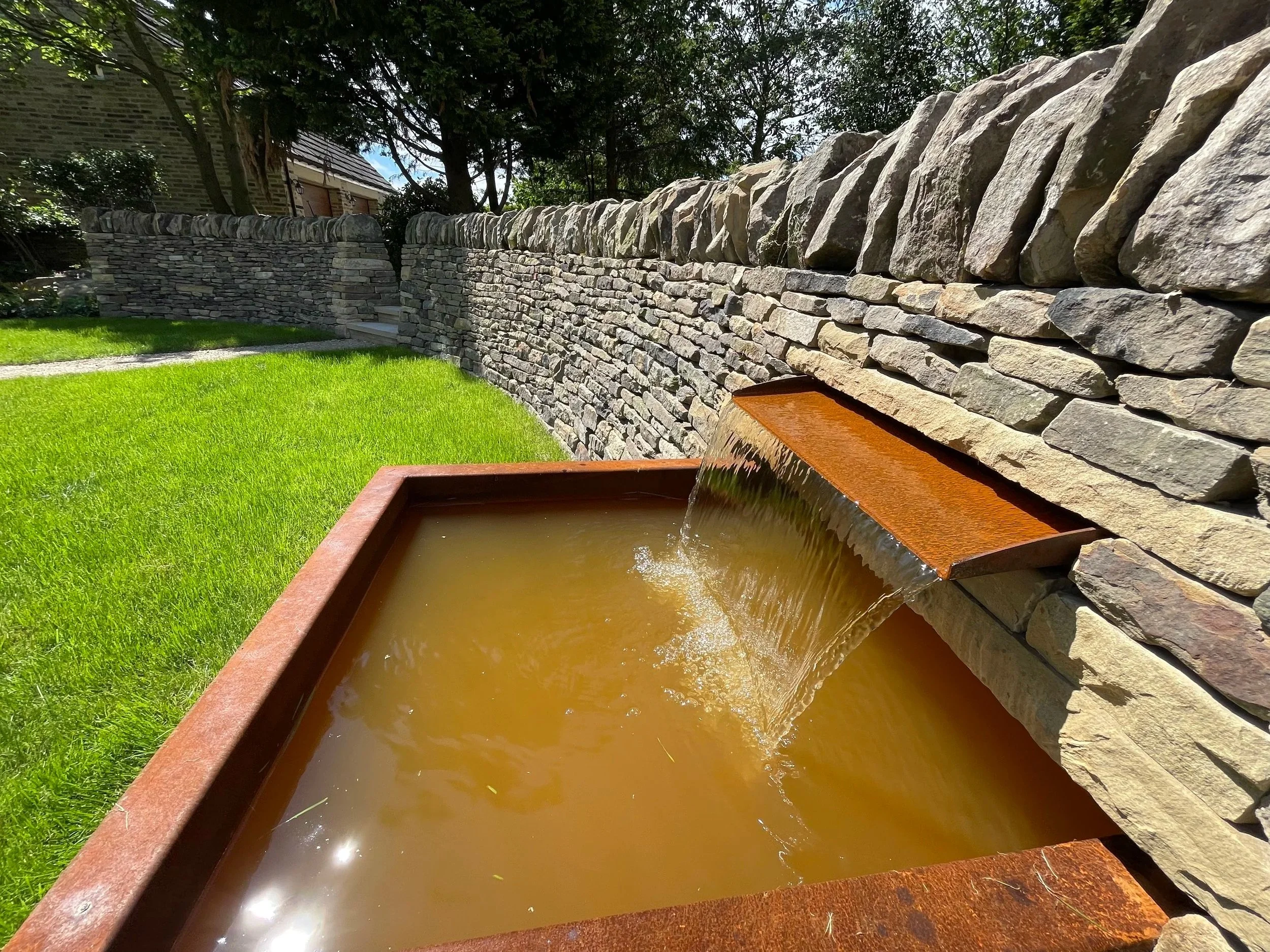 A stone wall with large rocks on top, green grass, and a water fountain feature at the bottom.