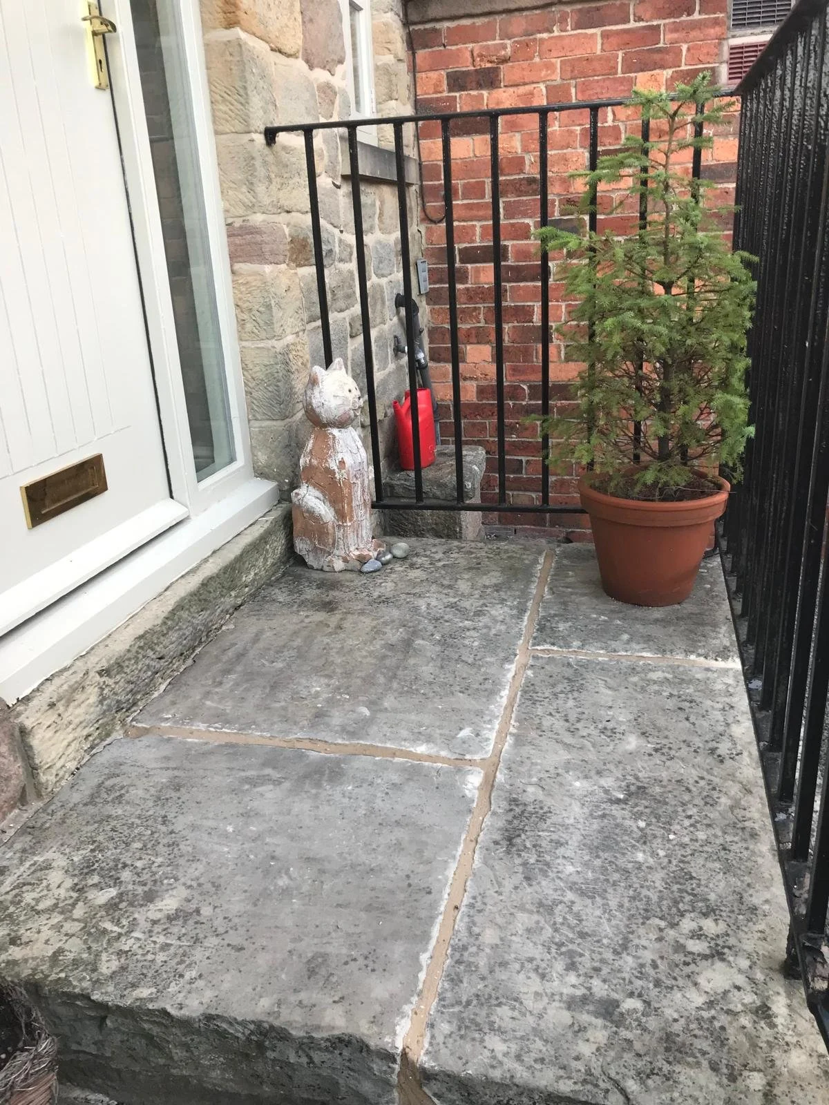 A small outdoor patio with large concrete slabs. There is a potted rosemary plant on the right, a small clay dog statue in the middle, and a red fire extinguisher behind the dog statue. The patio is enclosed by a black metal railing, and the left side has a stone wall and a white door.