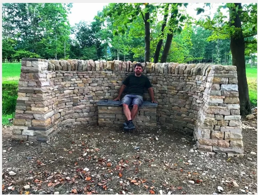 A man sitting on a stone bench with a tall stone wall behind him, in a park with green trees.