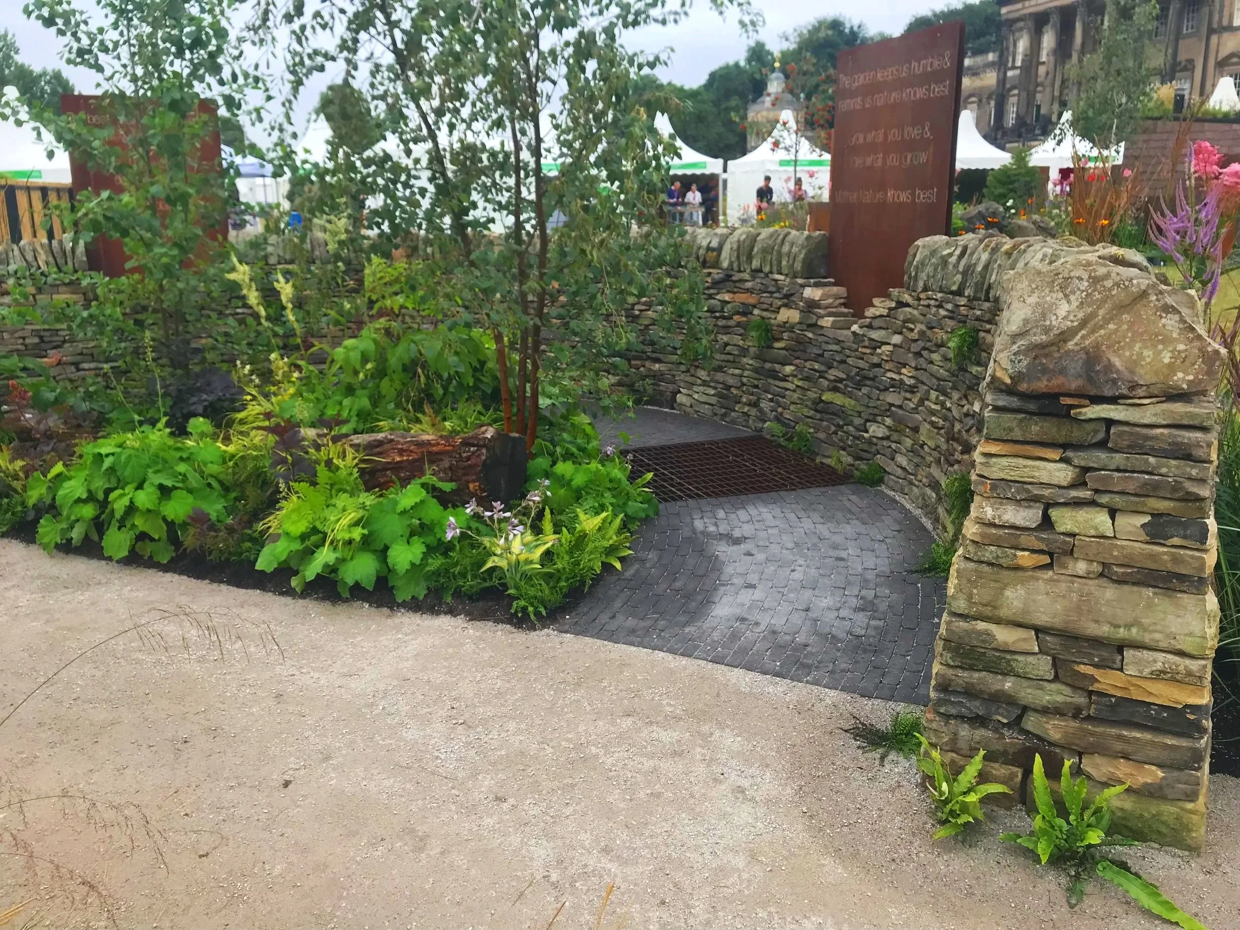 A garden with various green plants and flowers, a stone wall, and a paved pathway. In the background, there are white tents and people walking, with a historic building in the distance.