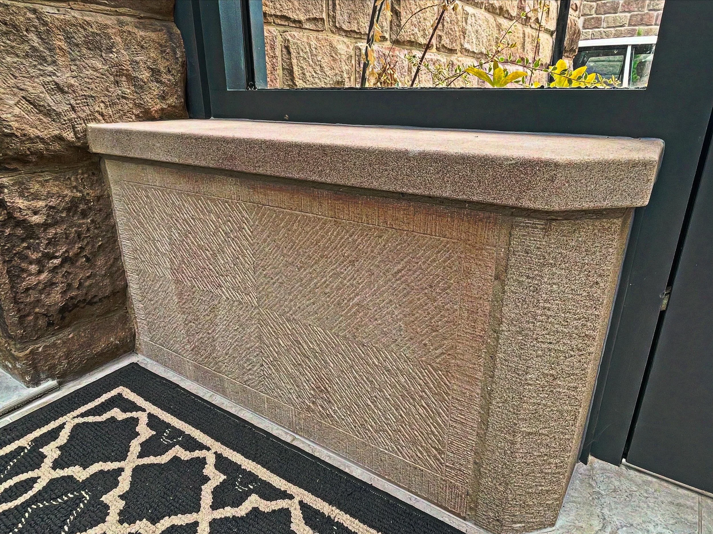 Close-up of a porch area with a textured stone wall and a black metal-framed glass door. In front, there is a beige stone ledge and a black and white patterned doormat on the floor.