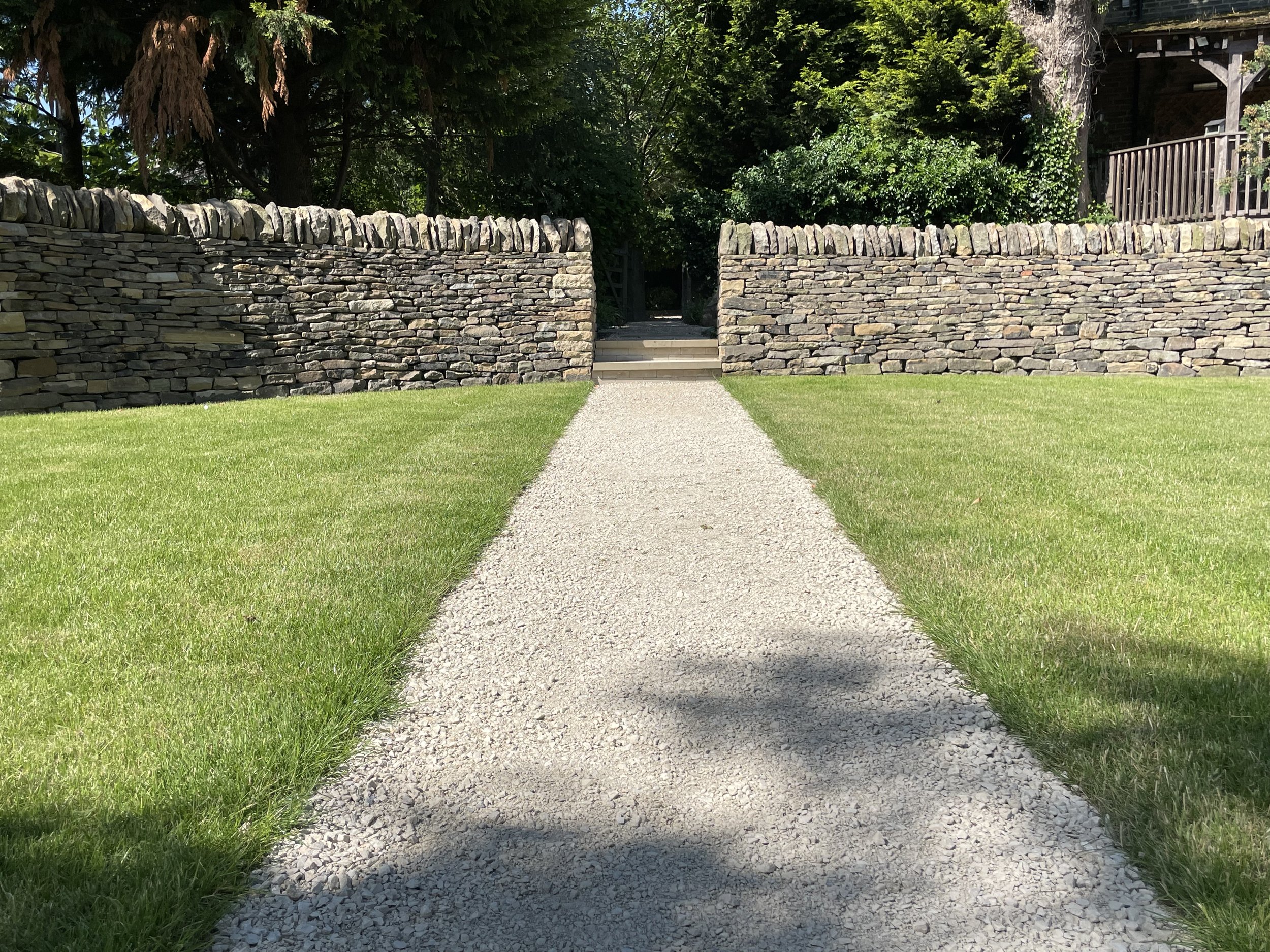 A gravel pathway leading through a stone wall with a small opening, green grass on either side, and trees and a wooden house in the background.